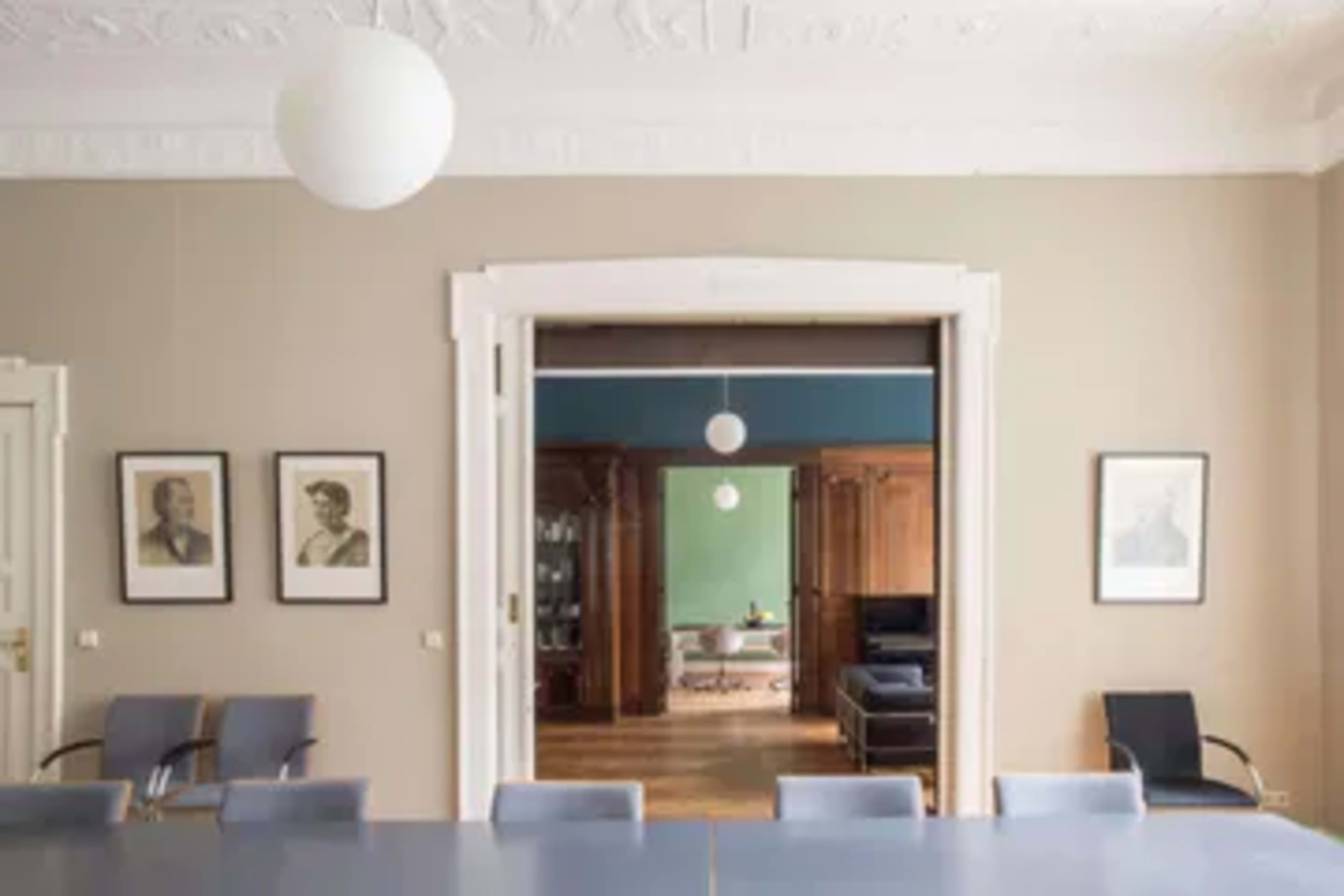 A spacious room with a long table and chairs, leading to another room featuring wooden furniture and green walls.