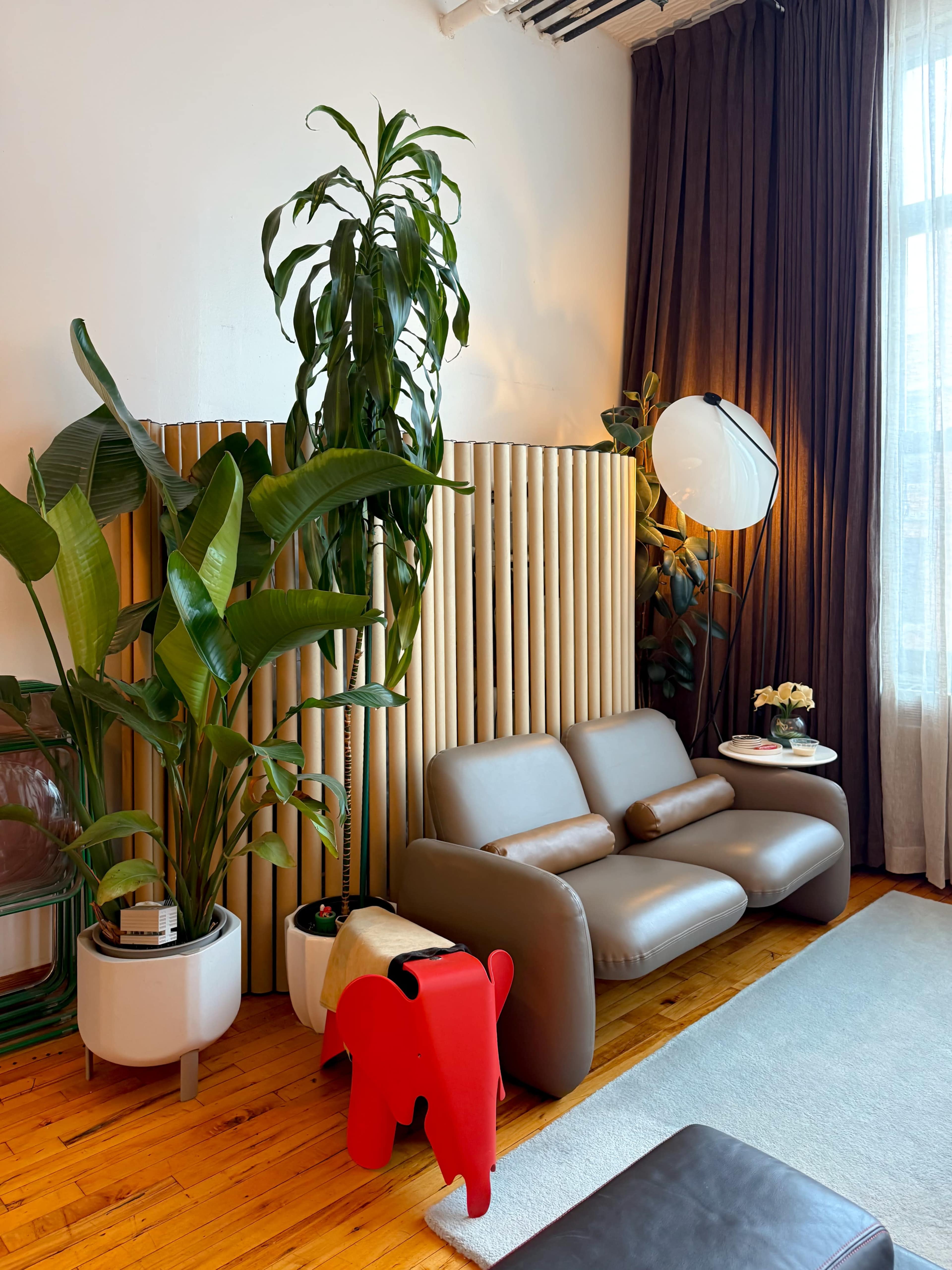 The image shows a modern living room with a light brown sofa, a red elephant-shaped stool, tall green plants, and a round lamp beside a textured wooden divider.