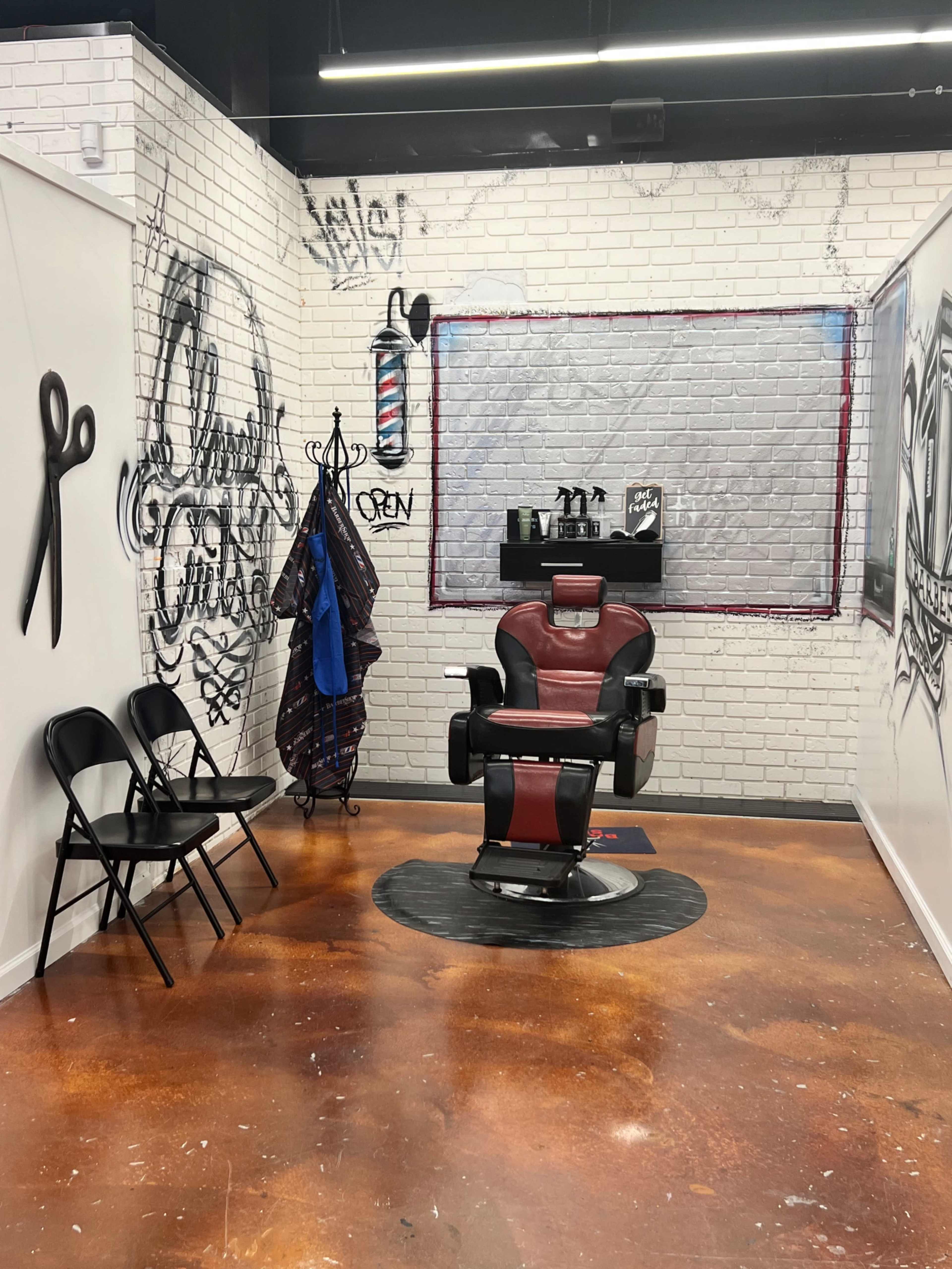 A modern barber shop interior with a red barber chair, a wall decorated with graffiti-style art, and a waiting area featuring black folding chairs.