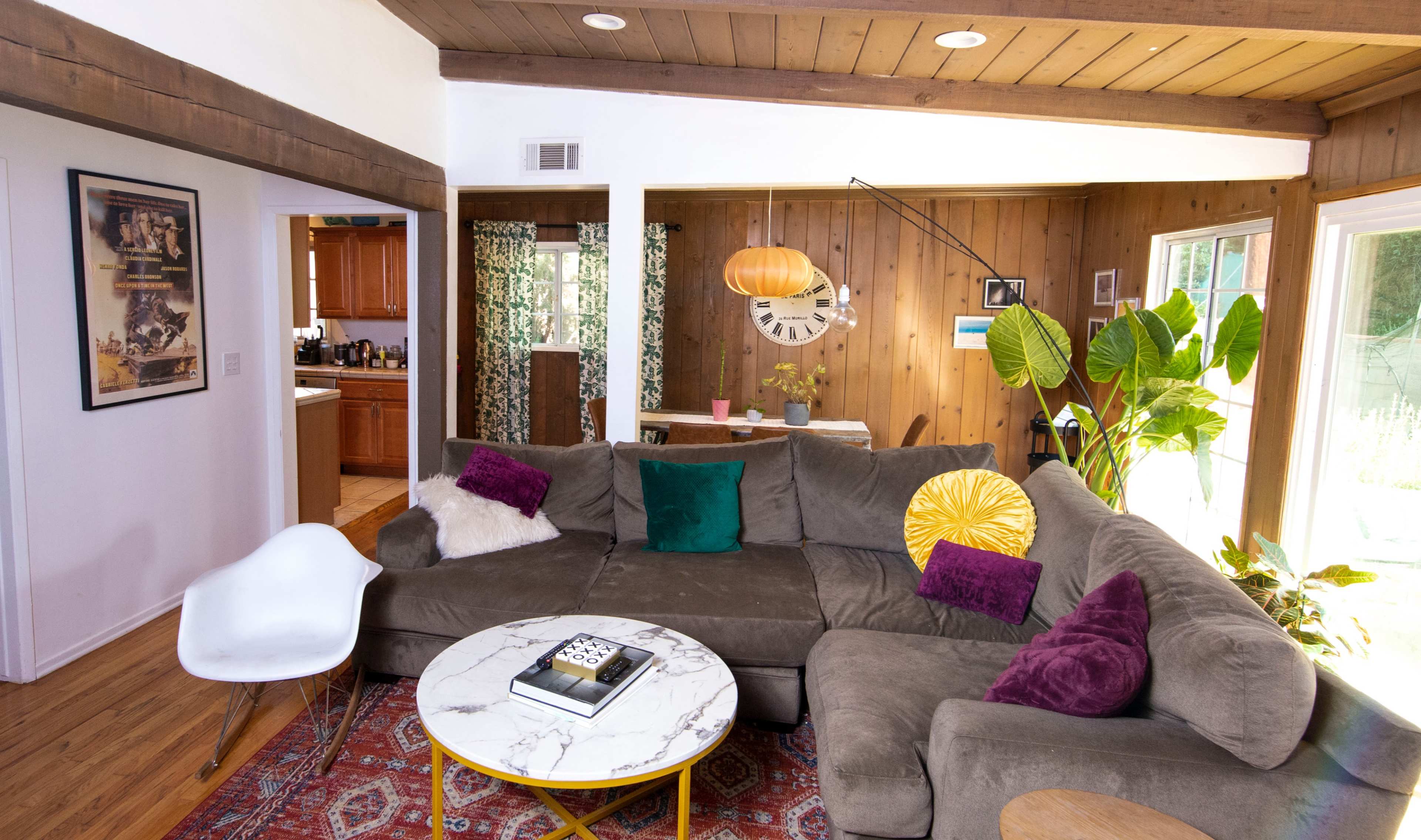 A spacious living room with a large sectional couch, a round coffee table, and a dining area in the background, adorned with plants and decorative elements.