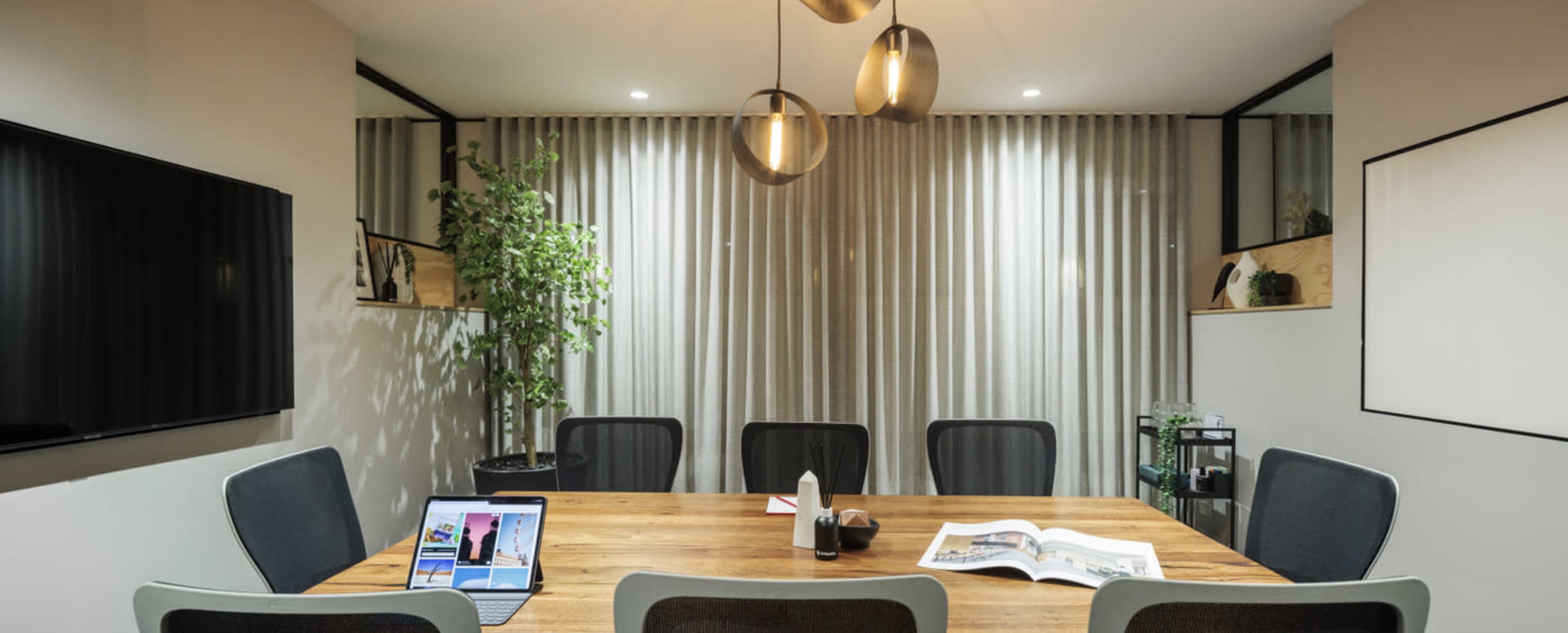 A modern conference room features a large wooden table surrounded by eight chairs, with a television on the wall and a plant in the corner.