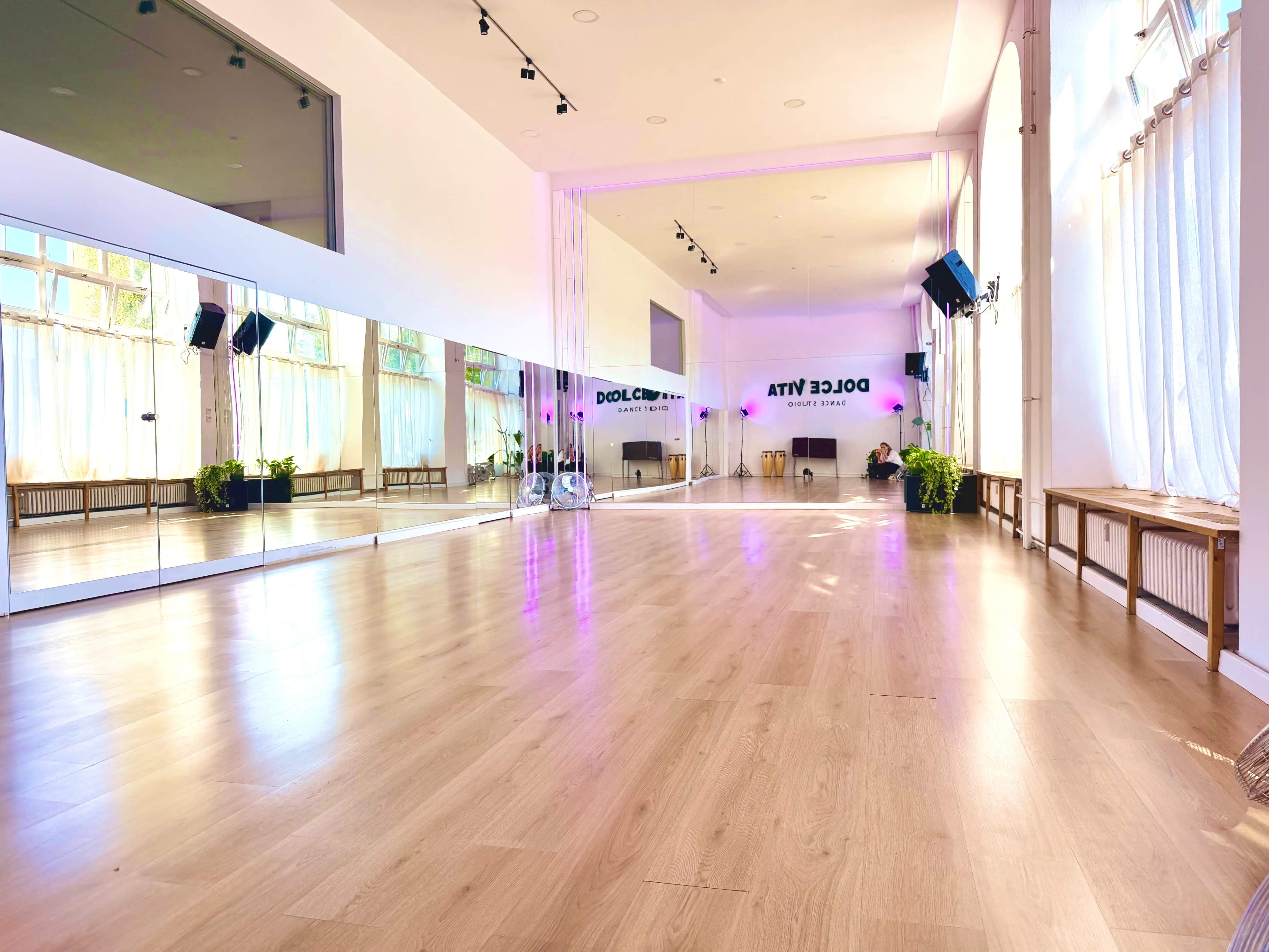 The image shows a bright dance studio with large mirrors, wooden flooring, and ample natural light streaming through white curtains.