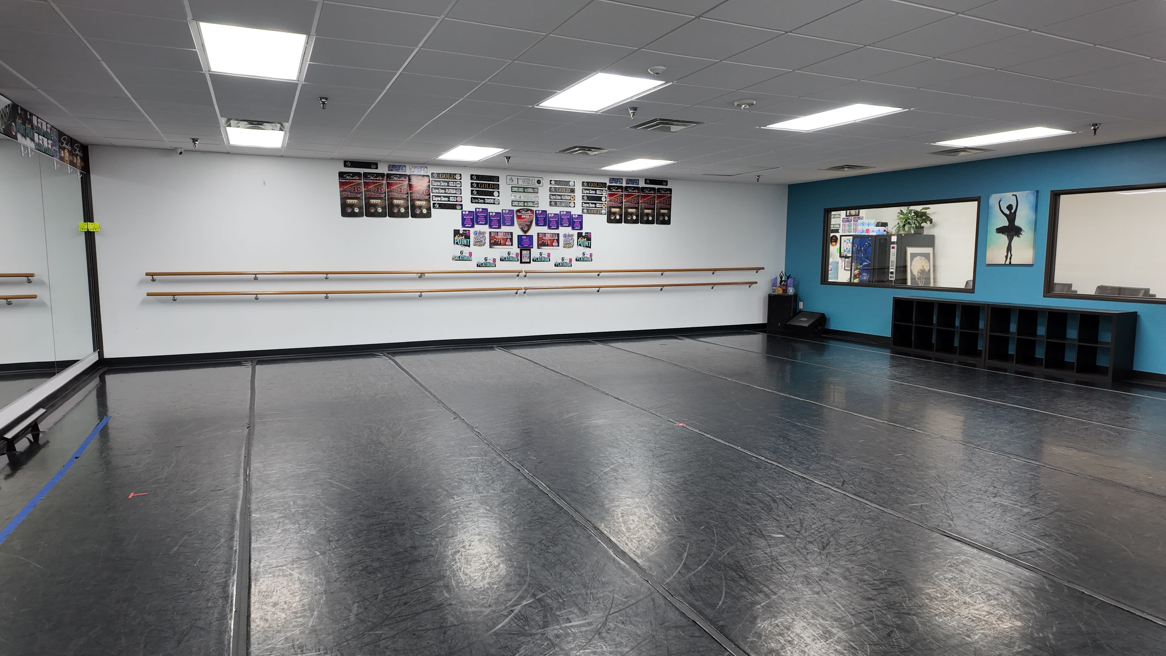 The image shows a spacious dance studio with a mirror wall, ballet barres, and motivational posters on the walls.