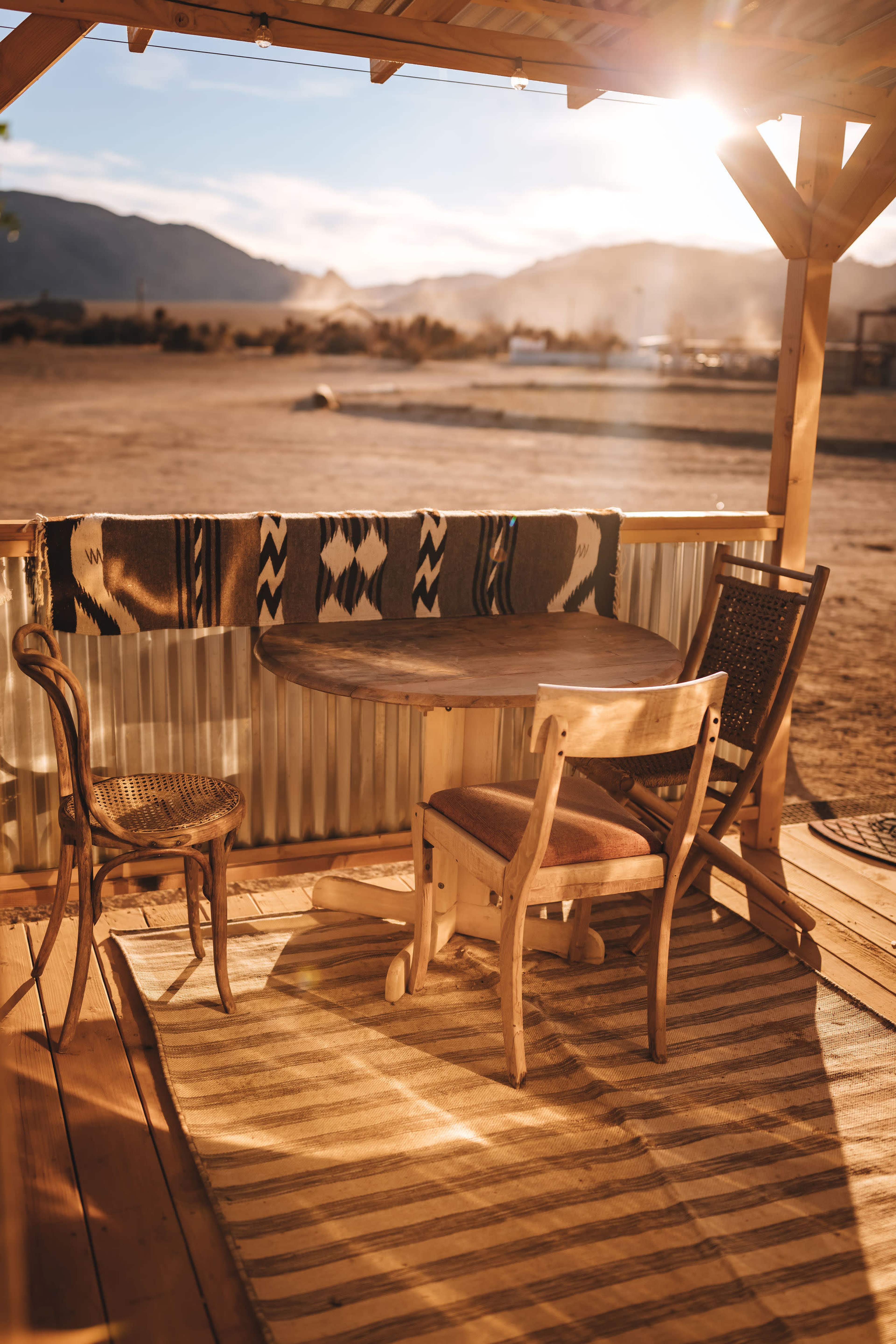 A wooden table and two chairs are positioned under a shelter with a blanket draped on the railing, set against a backdrop of mountains and a sunset.