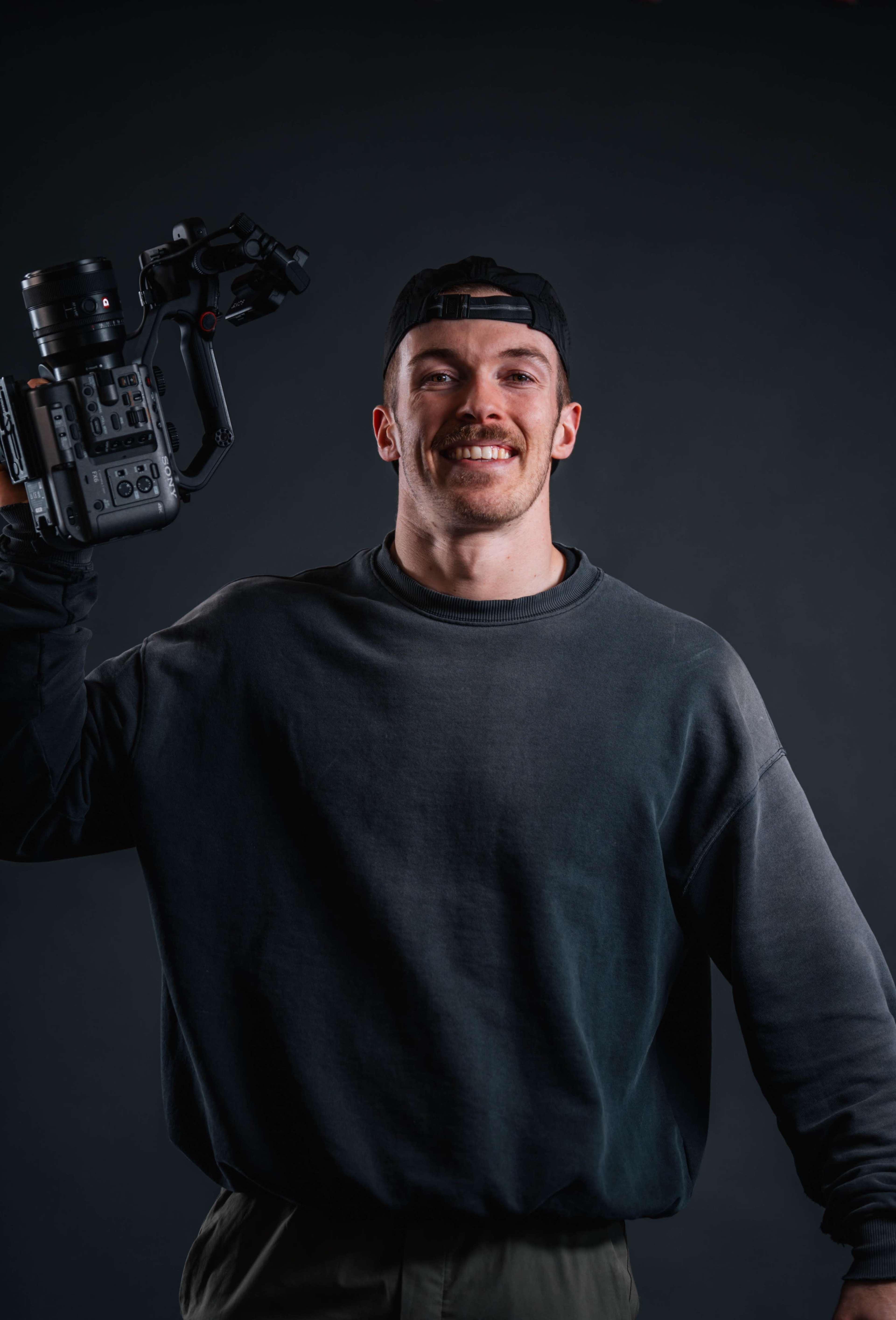 A man wearing a dark sweatshirt and cap holds a camera rig while smiling against a dark background.