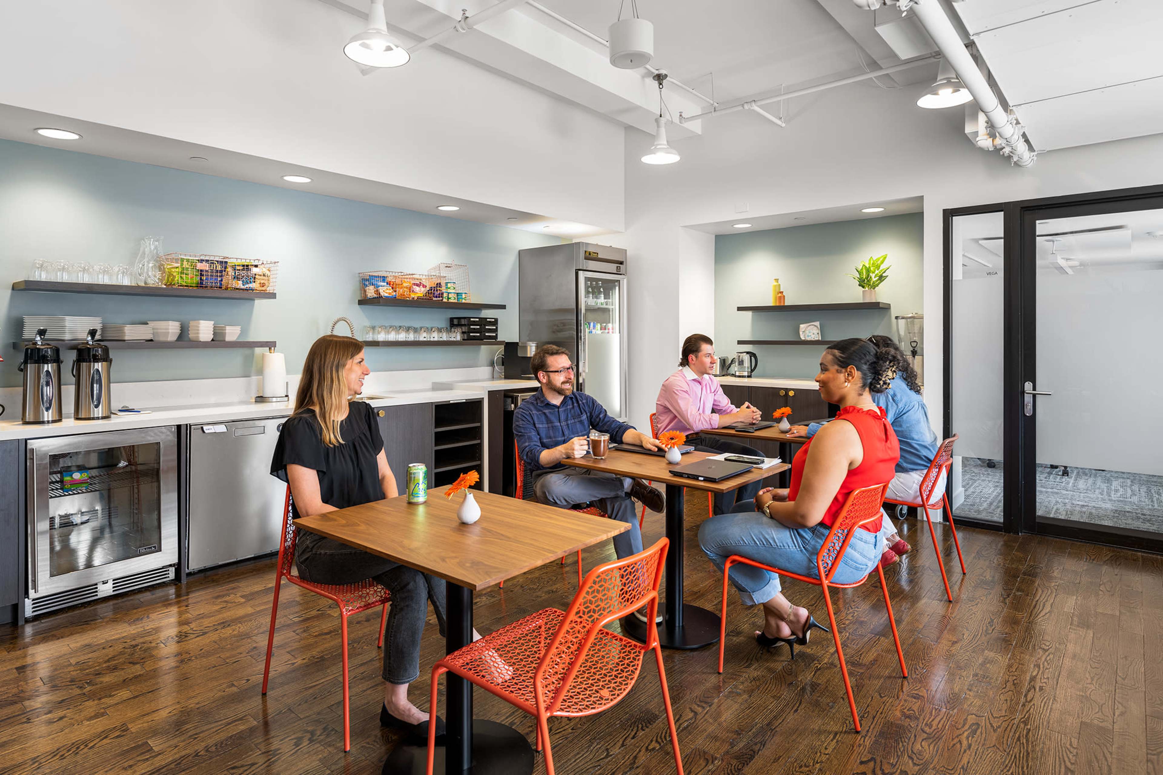 A group of five people is seated around tables in a modern break room with snacks and beverages available in the background.