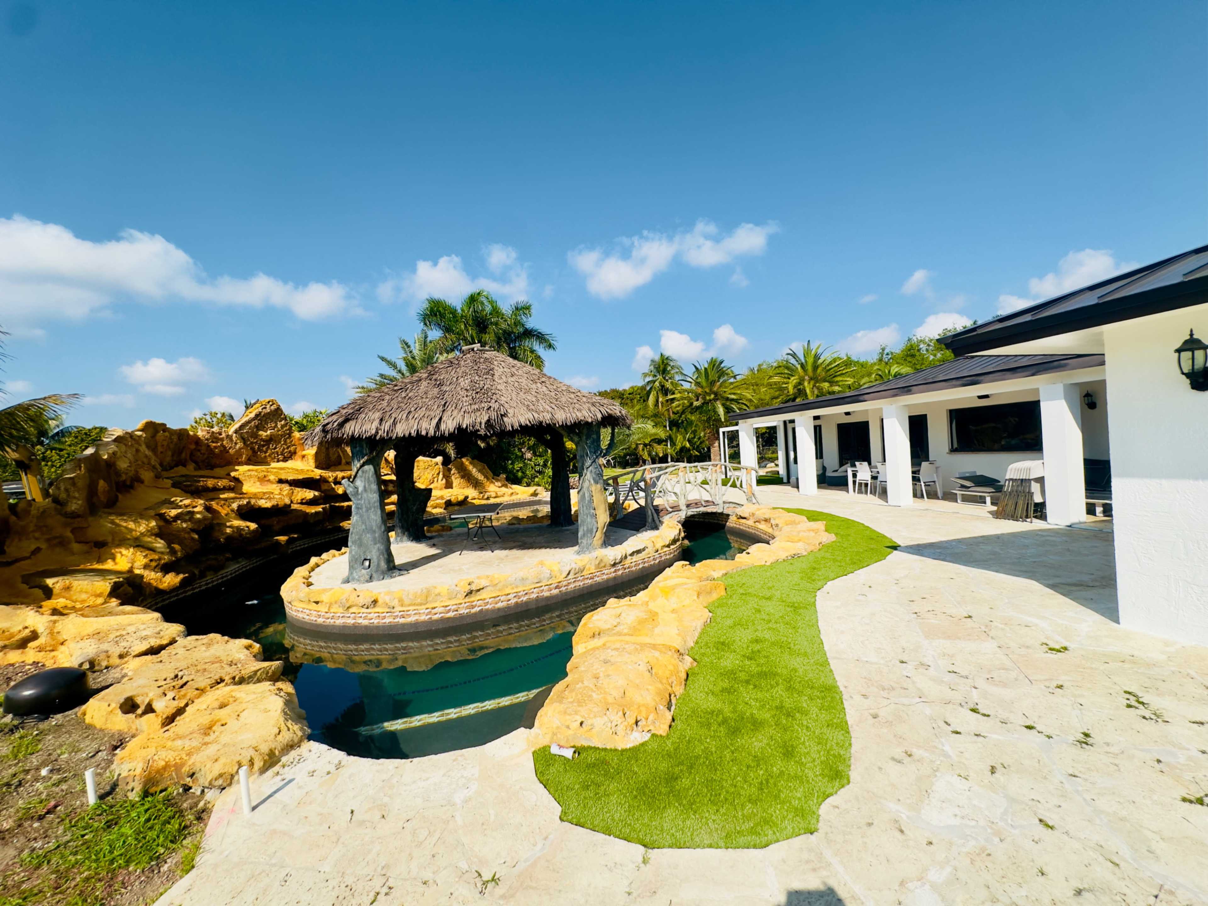 A thatched-roof gazebo overlooks a landscaped area with stone formations and a pool, adjacent to a white building.