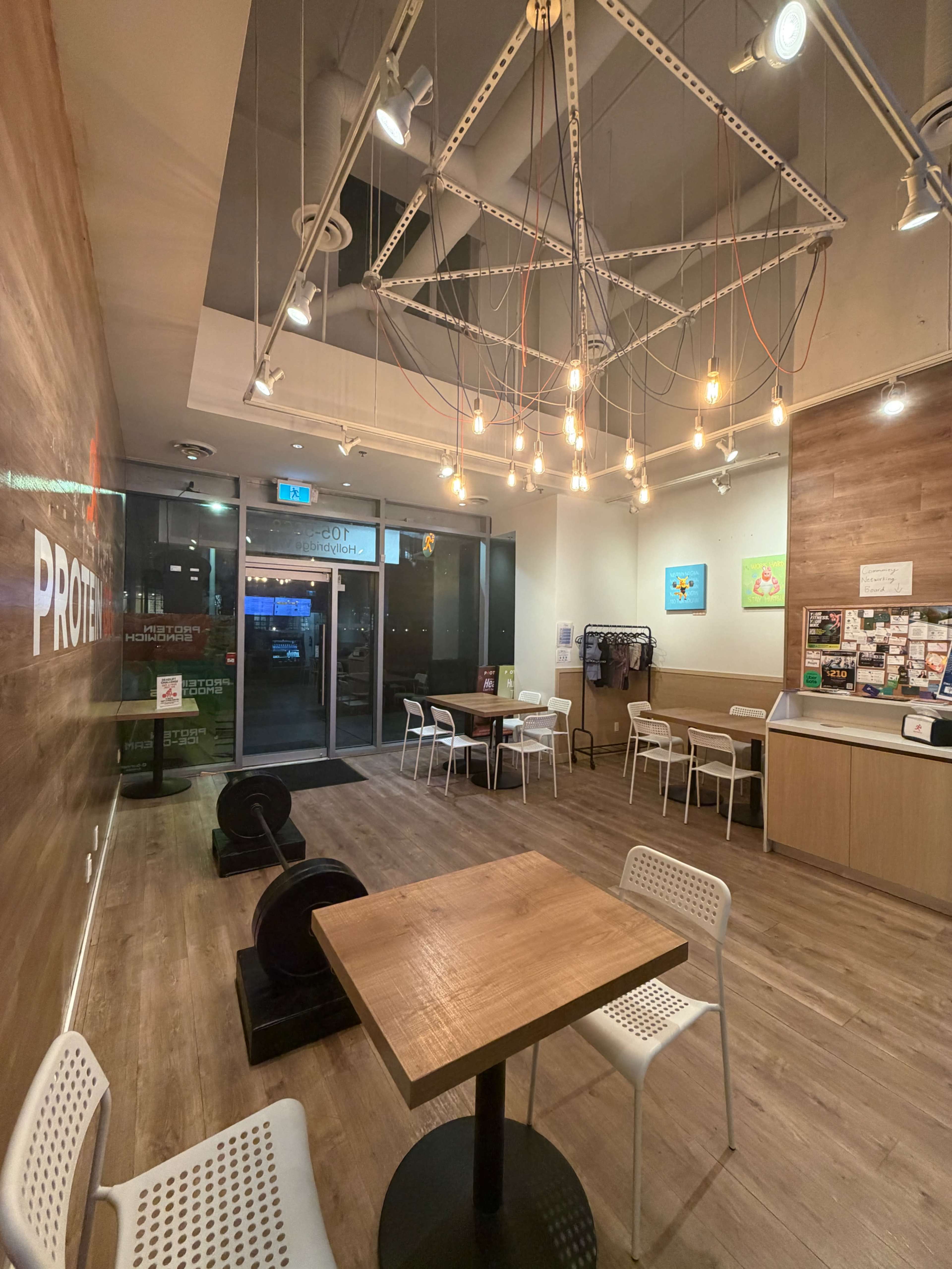 The image shows a modern, minimalistic café interior with wooden accents, featuring a large table and several white chairs, alongside a glass entrance door and wall decorations.
