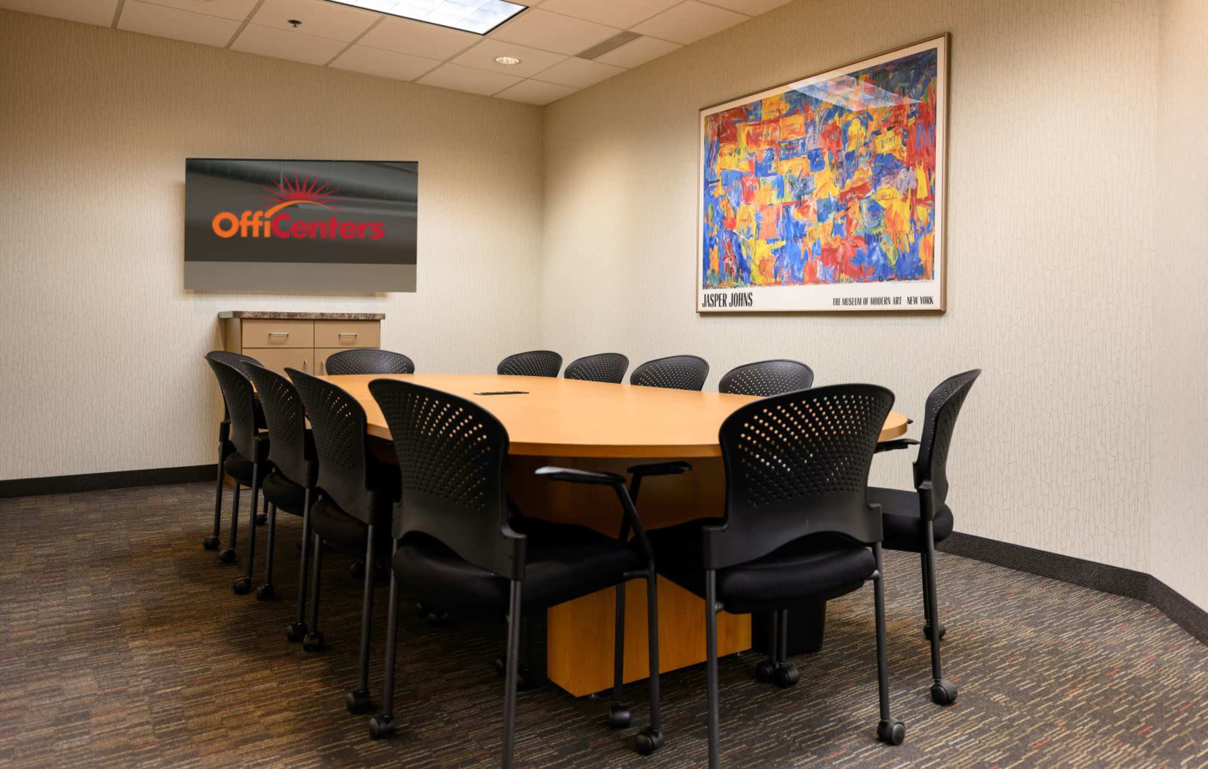 A conference room features a large oval table surrounded by black chairs and a wall-mounted screen displaying the logo "OffiCenters" alongside an abstract artwork.