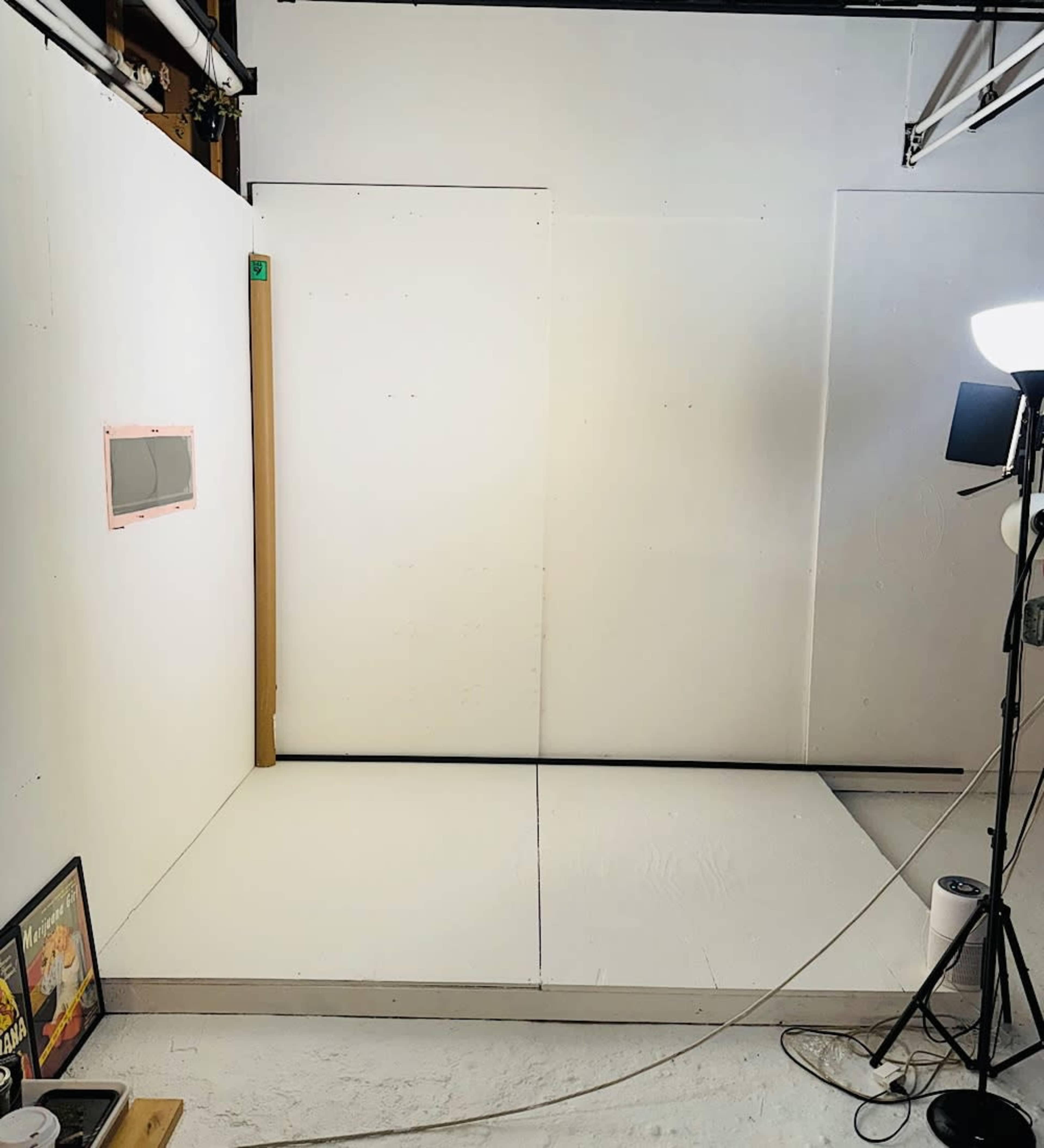 The image shows a minimalistic studio space with white walls, a small window, and lighting equipment set up on one side.