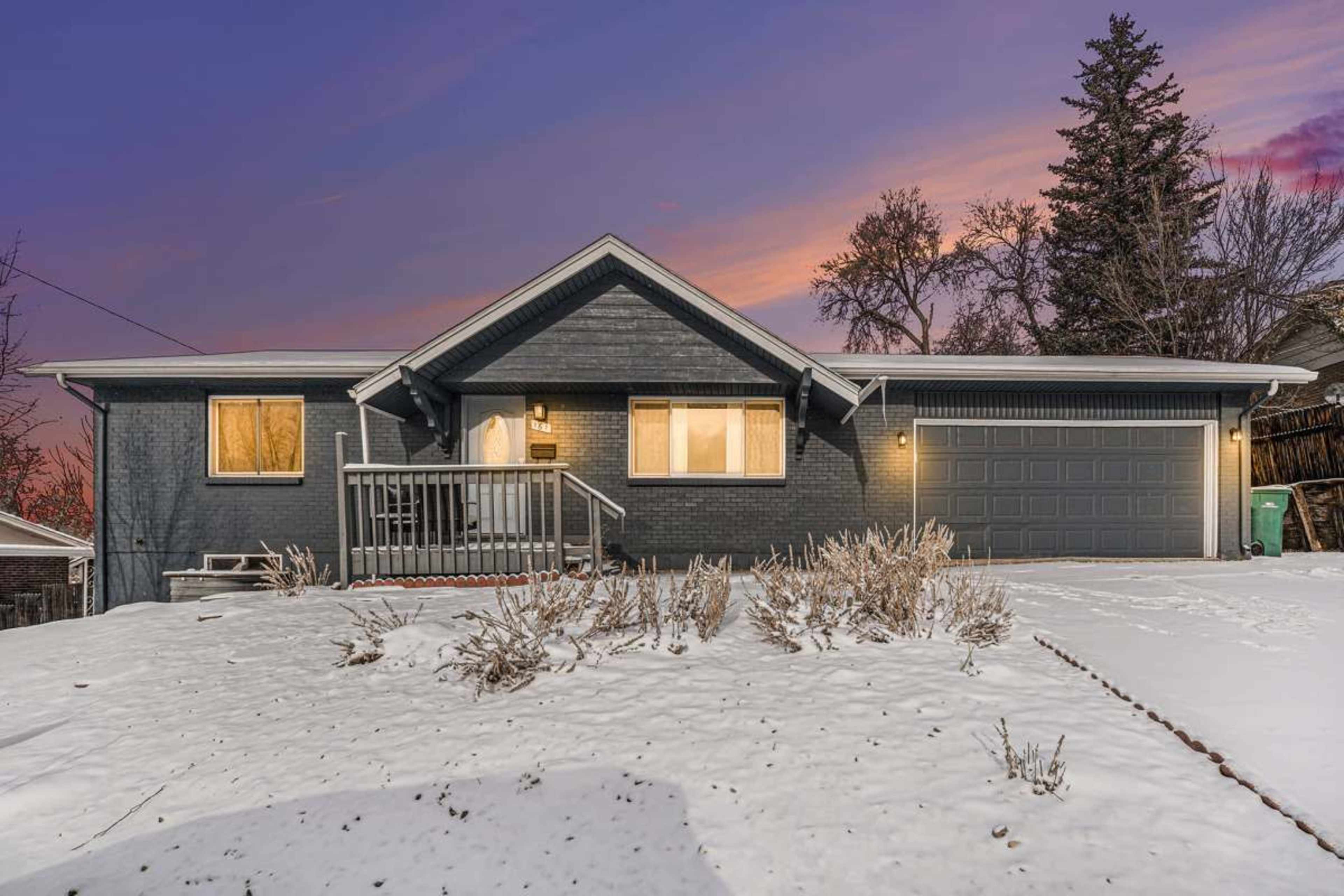 A single-story house with a gray exterior sits in a snowy yard at dusk, illuminated by exterior lighting.