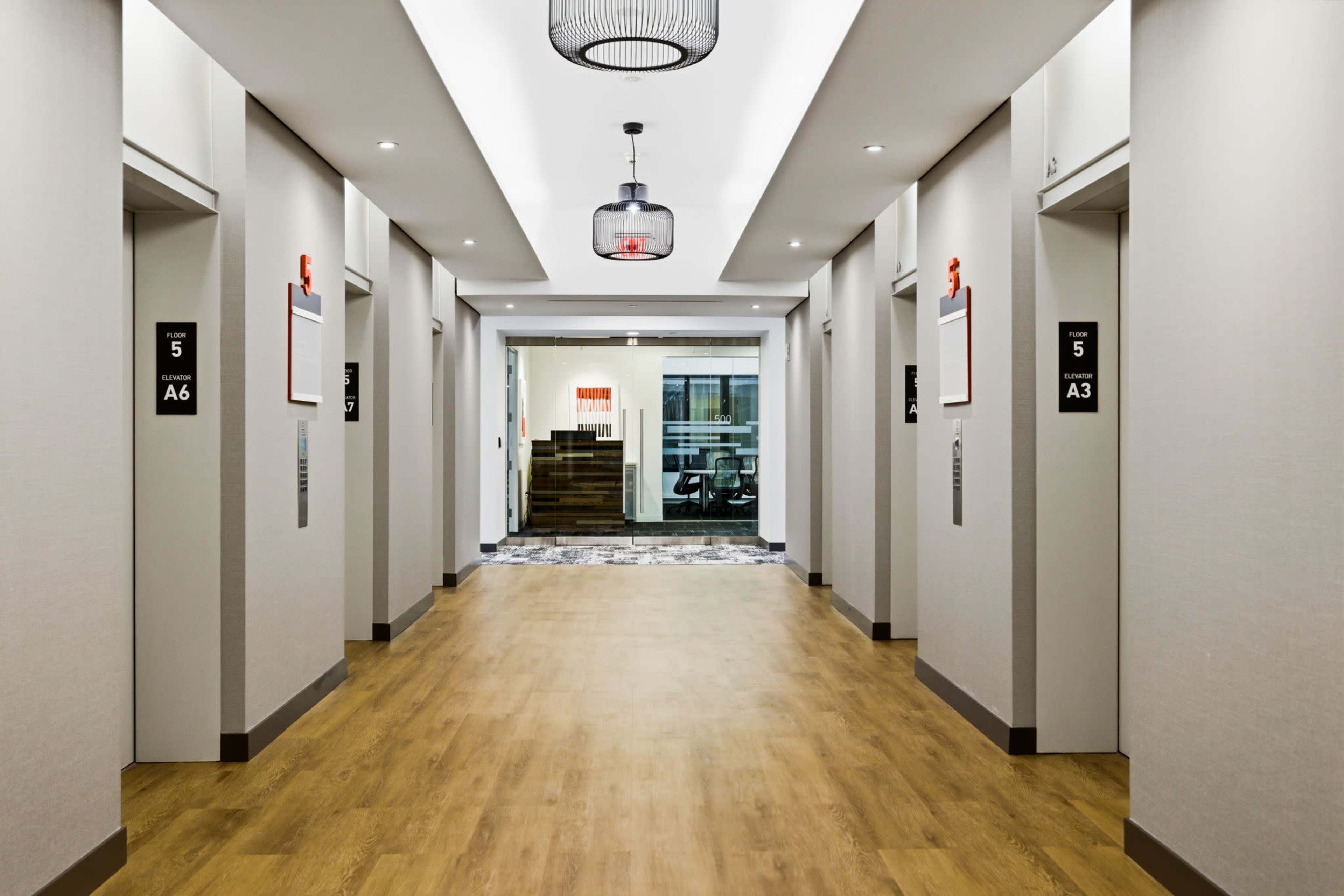 The image shows a well-lit hallway with several elevator doors on either side and a glass entrance at the end.