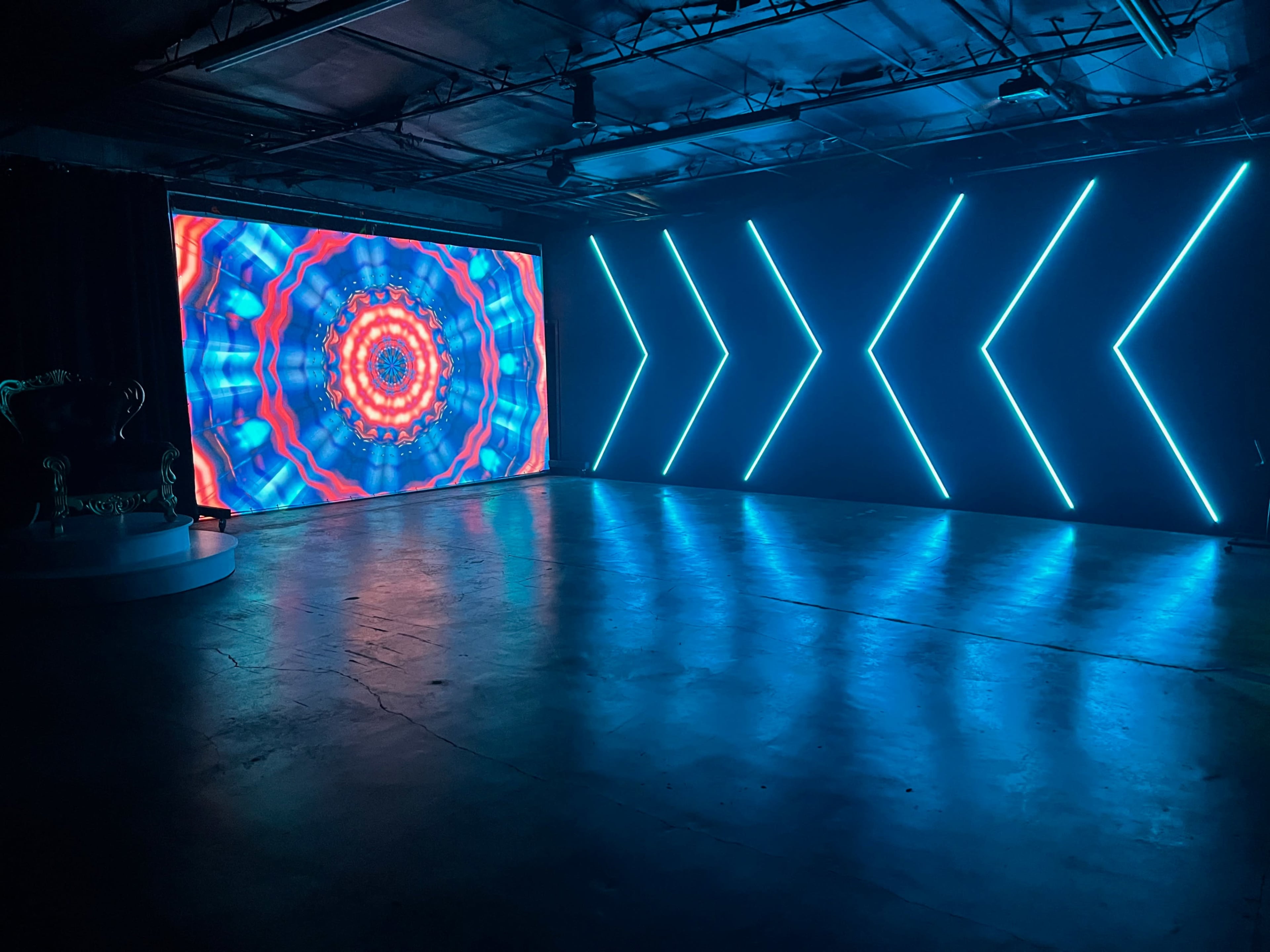 A brightly lit room features a large LED screen displaying a colorful kaleidoscopic pattern and a wall with neon blue arrow lighting.