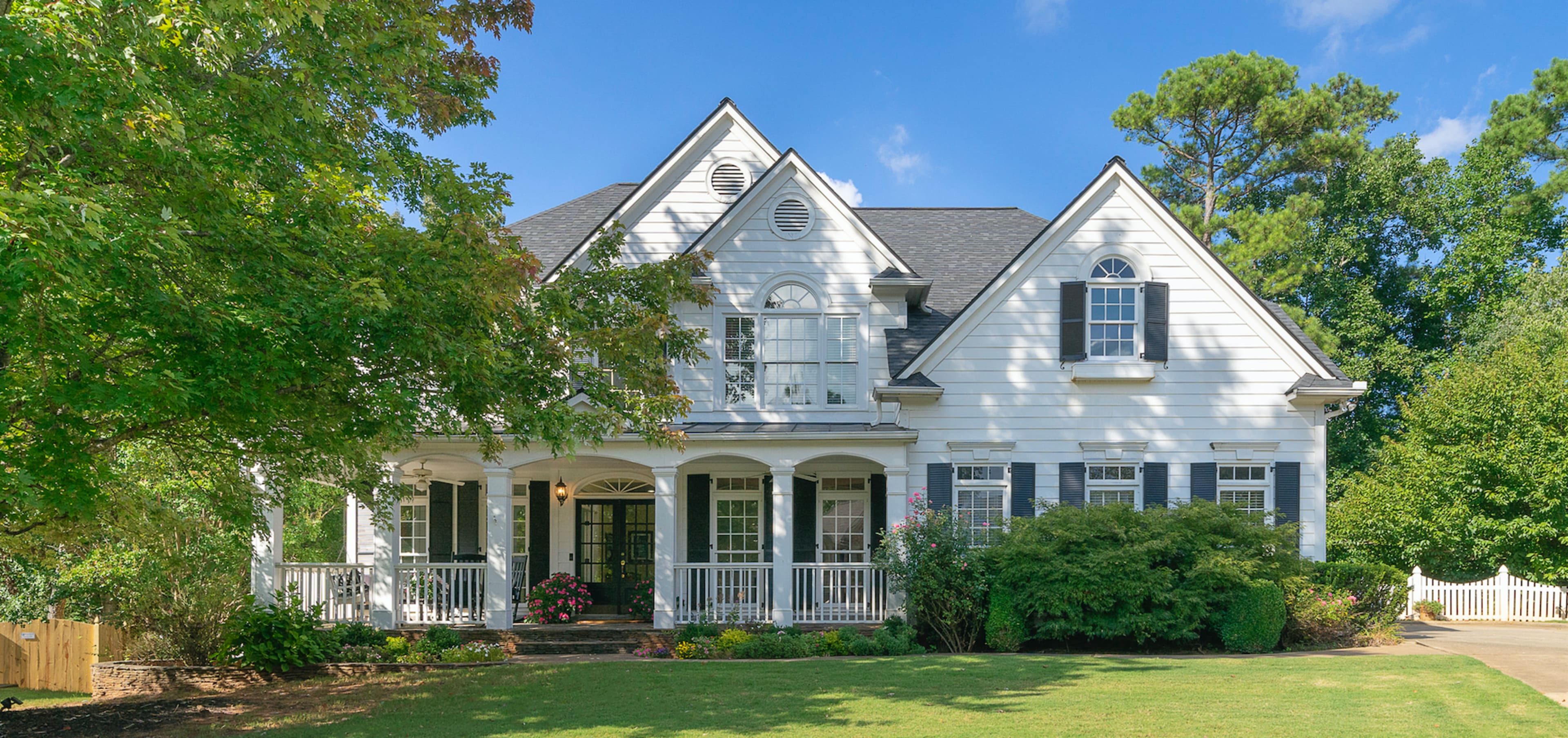 A large, two-story white house with a front porch is surrounded by greenery and has multiple windows.