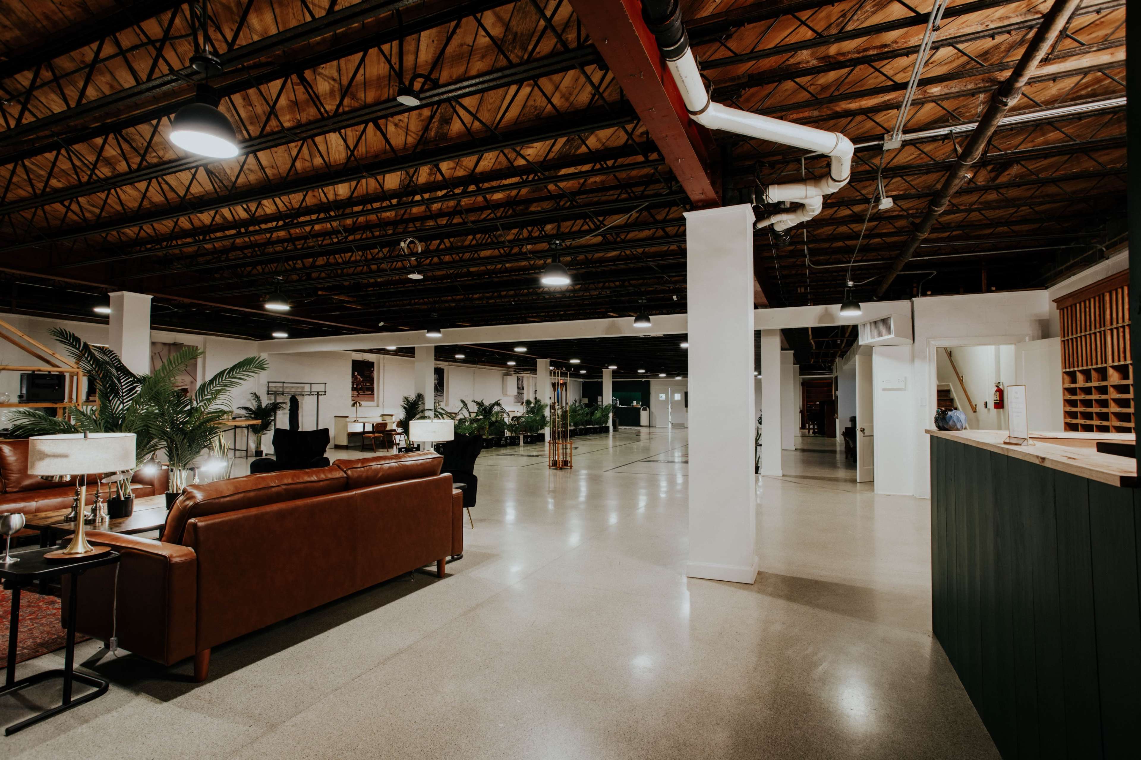 The image shows a spacious, modern interior with polished concrete floors, exposed beams, and a seating area featuring brown sofas and potted plants.