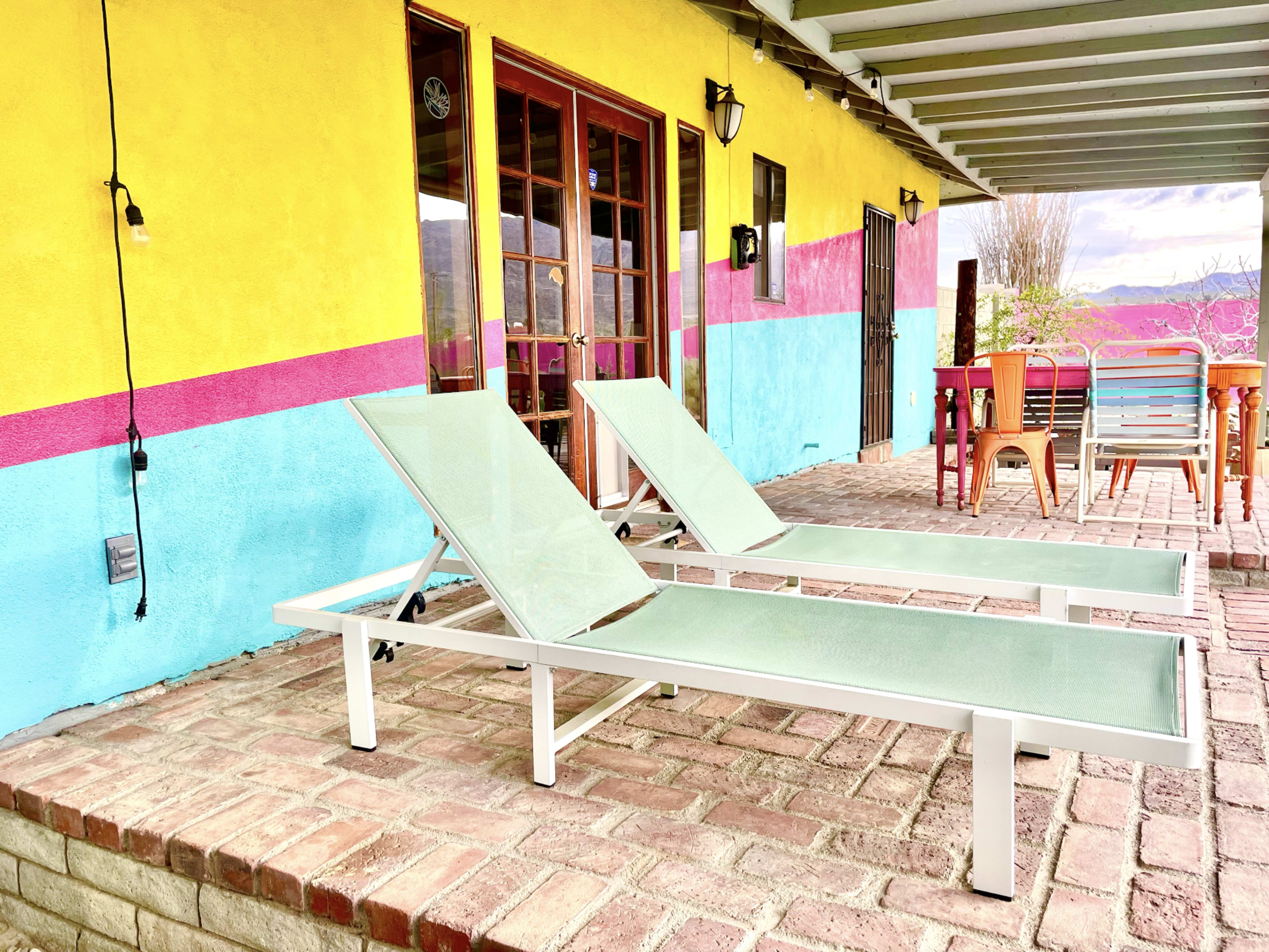 Two light green lounge chairs are positioned on a brick patio in front of a brightly painted house with yellow and turquoise walls and pink accents.