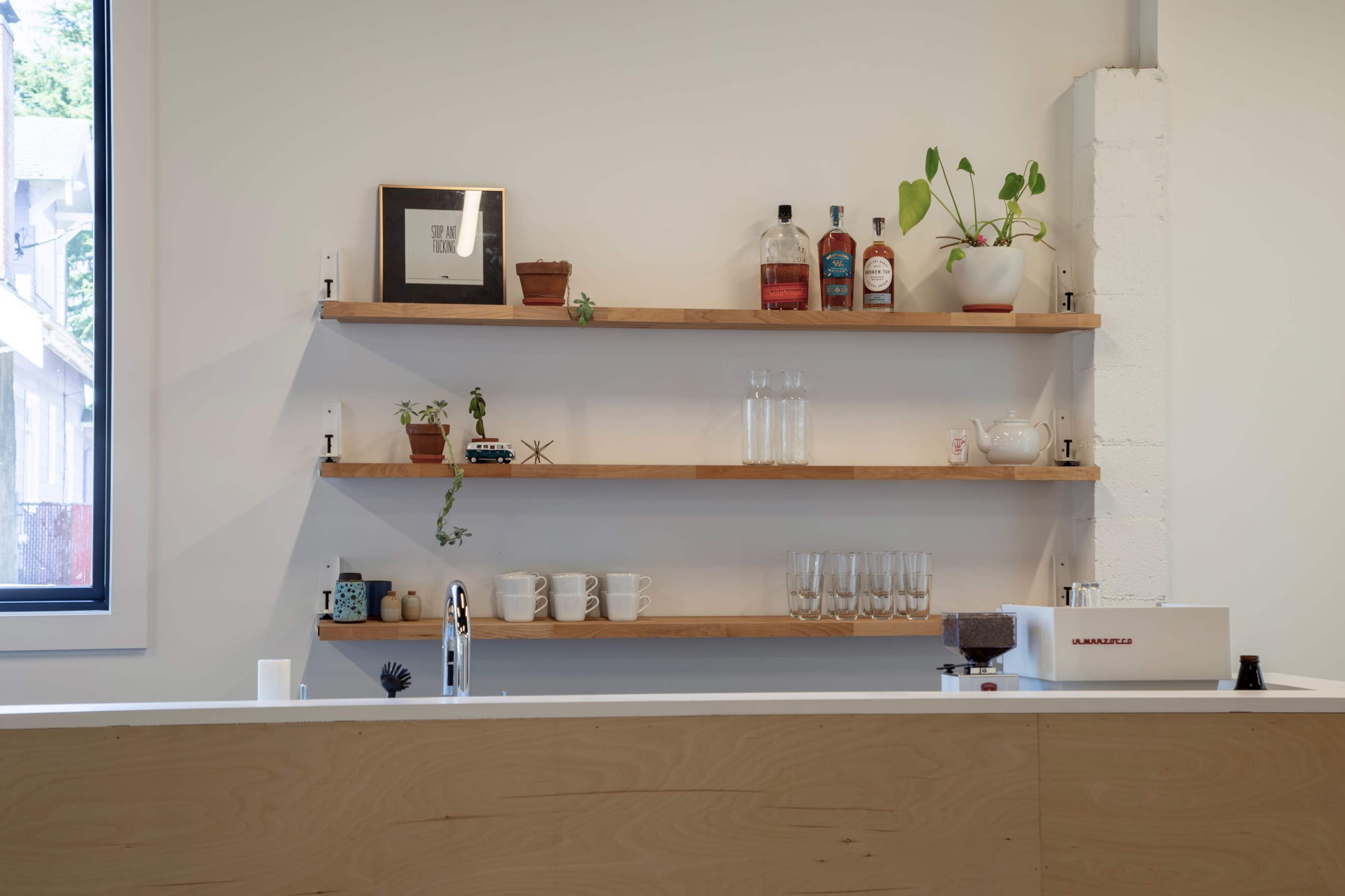 A minimalist kitchen counter with wooden shelves displaying various bottles, plants, and glassware against a plain wall.