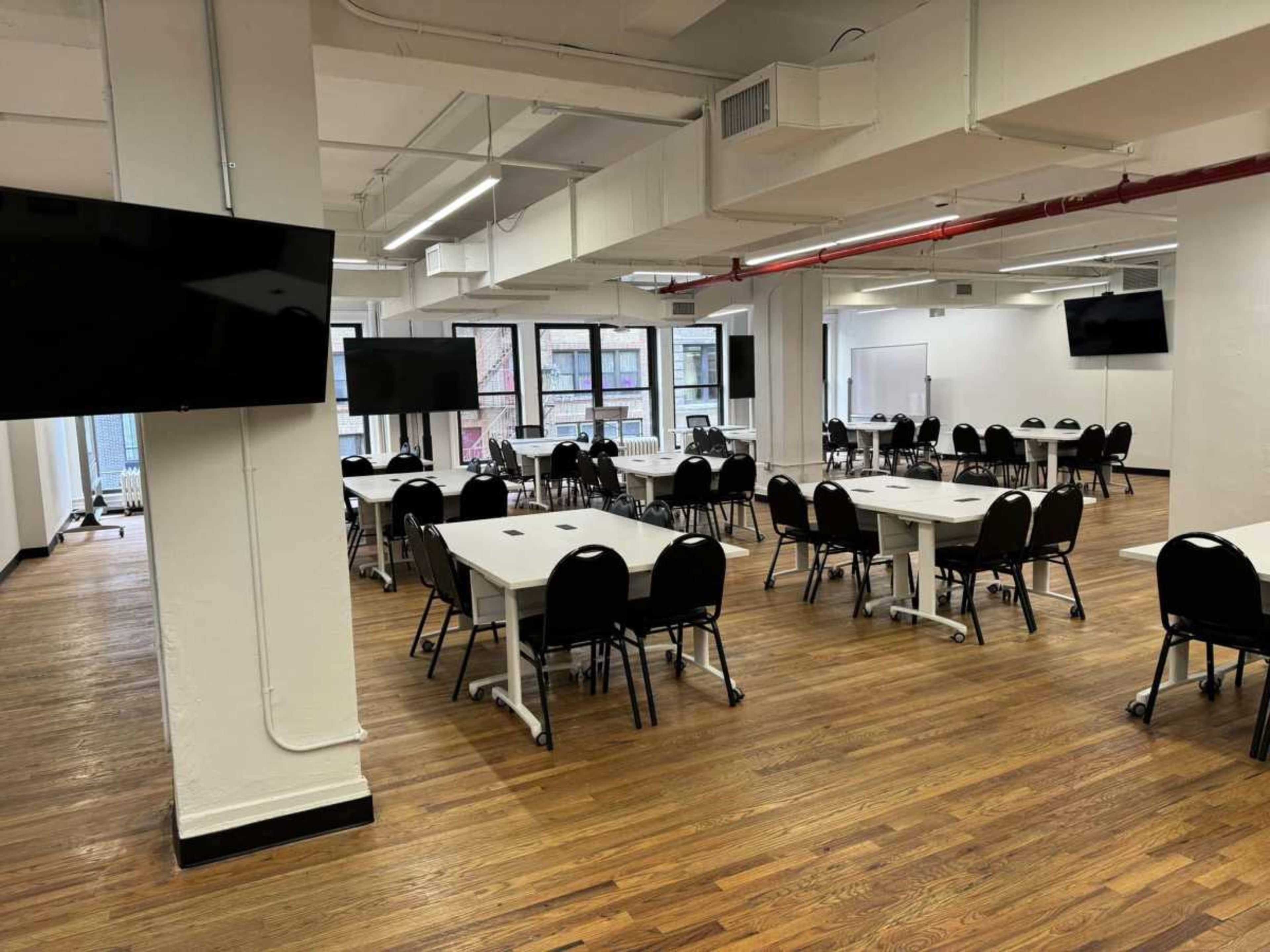 The image shows a spacious, well-lit room arranged with multiple tables and chairs, along with two large screens mounted on the walls.