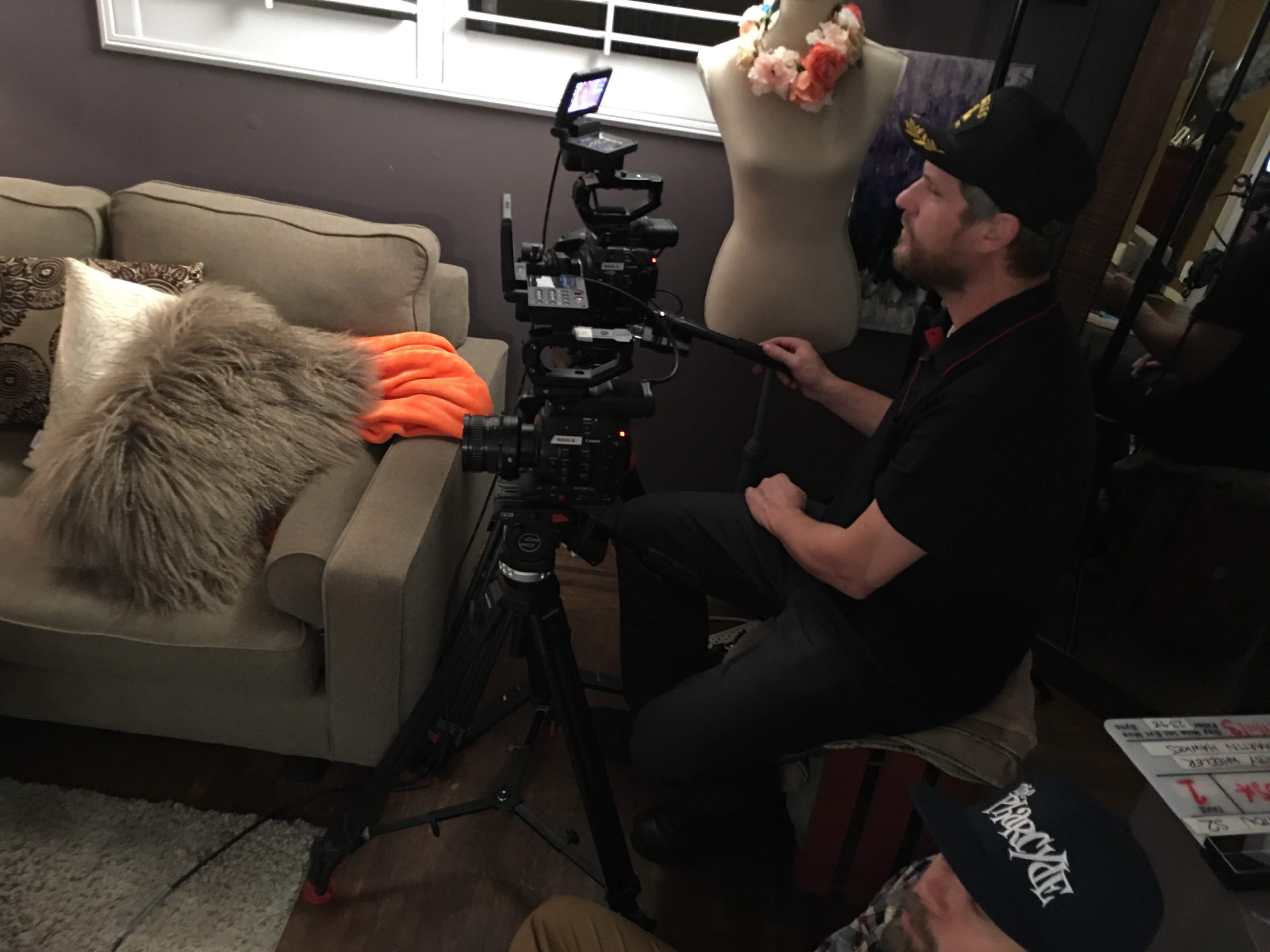 A cameraman operates a professional camera on a tripod while sitting on a stool in a room with a couch and a dress form.