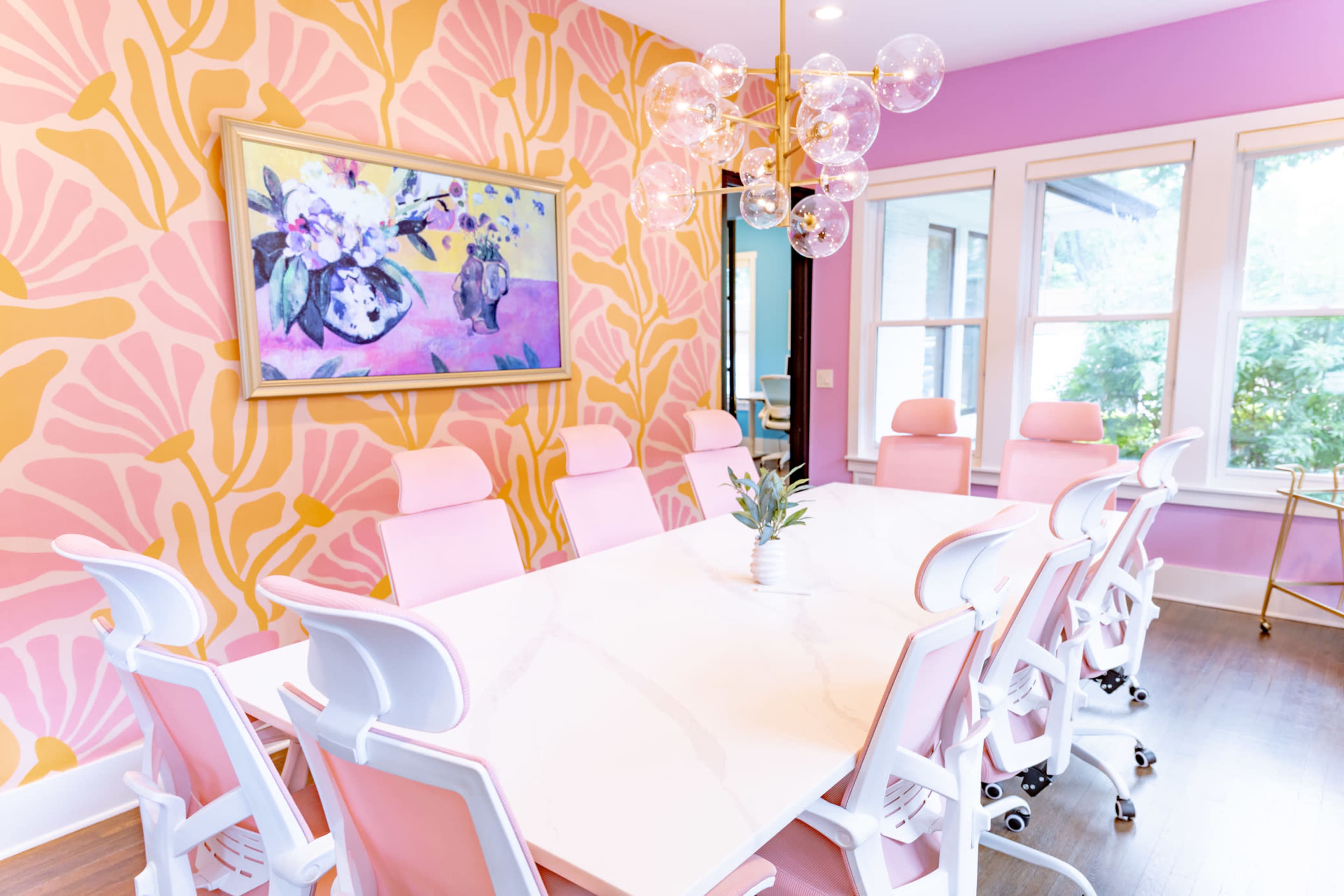 The image depicts a brightly colored conference room with a large marble table surrounded by pink office chairs and vibrant floral wallpaper.