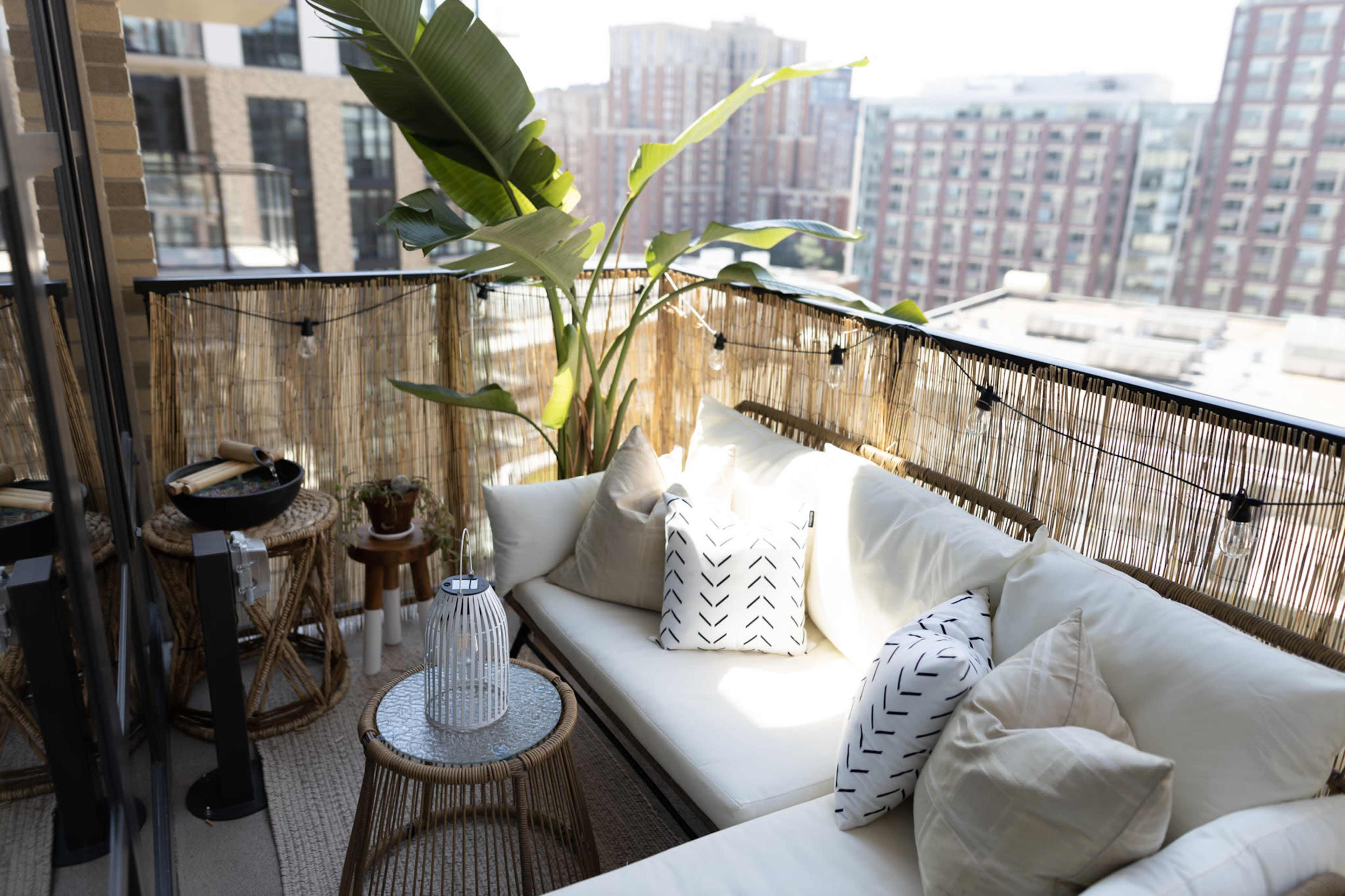 A modern balcony features a light-colored sofa adorned with patterned pillows, a round coffee table, and potted greenery, all surrounded by a bamboo screening and string lights.