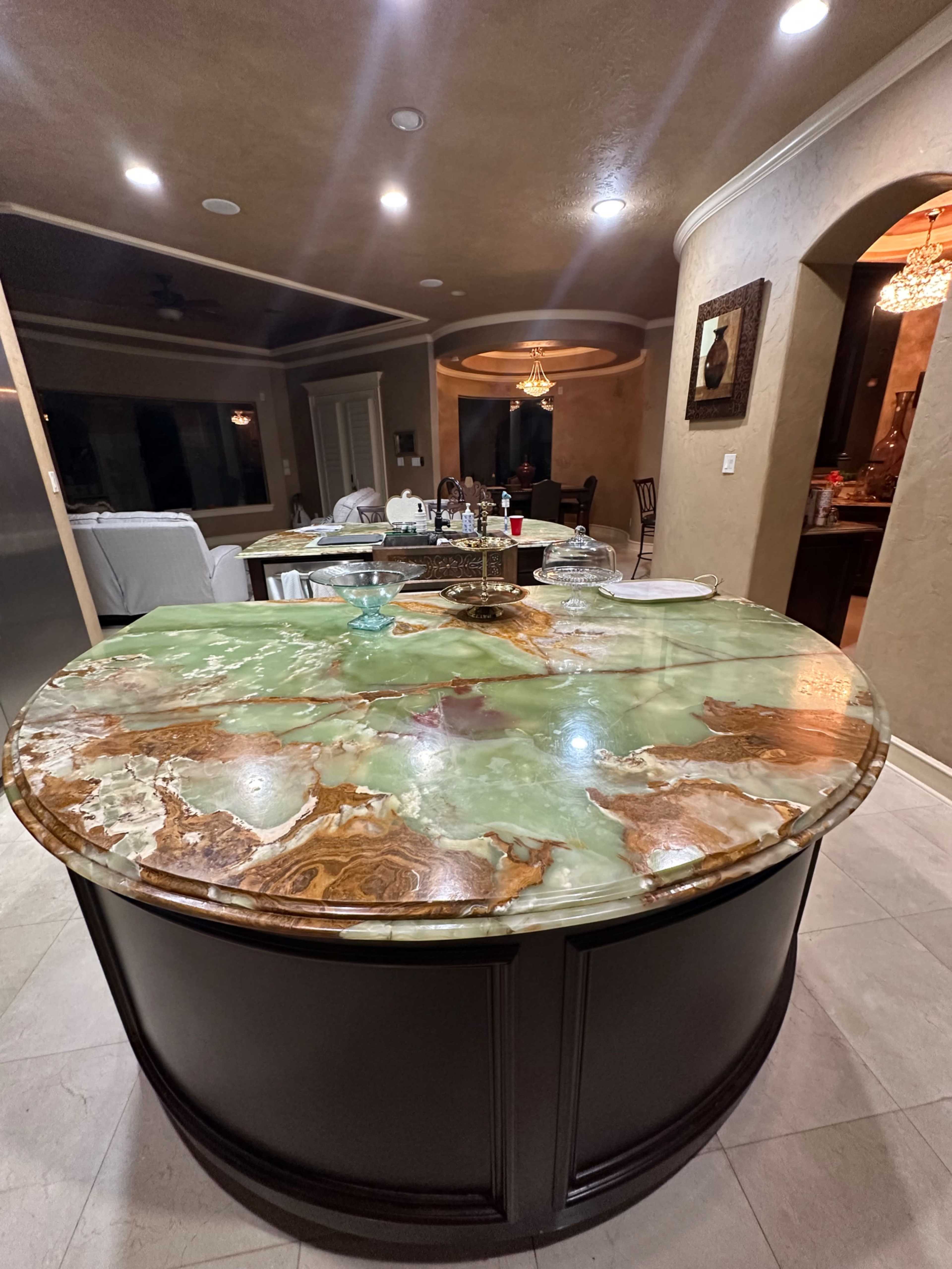 The image showcases a large, round kitchen island with a polished, multicolored stone countertop and elegant cabinetry, illuminated by recessed lighting.
