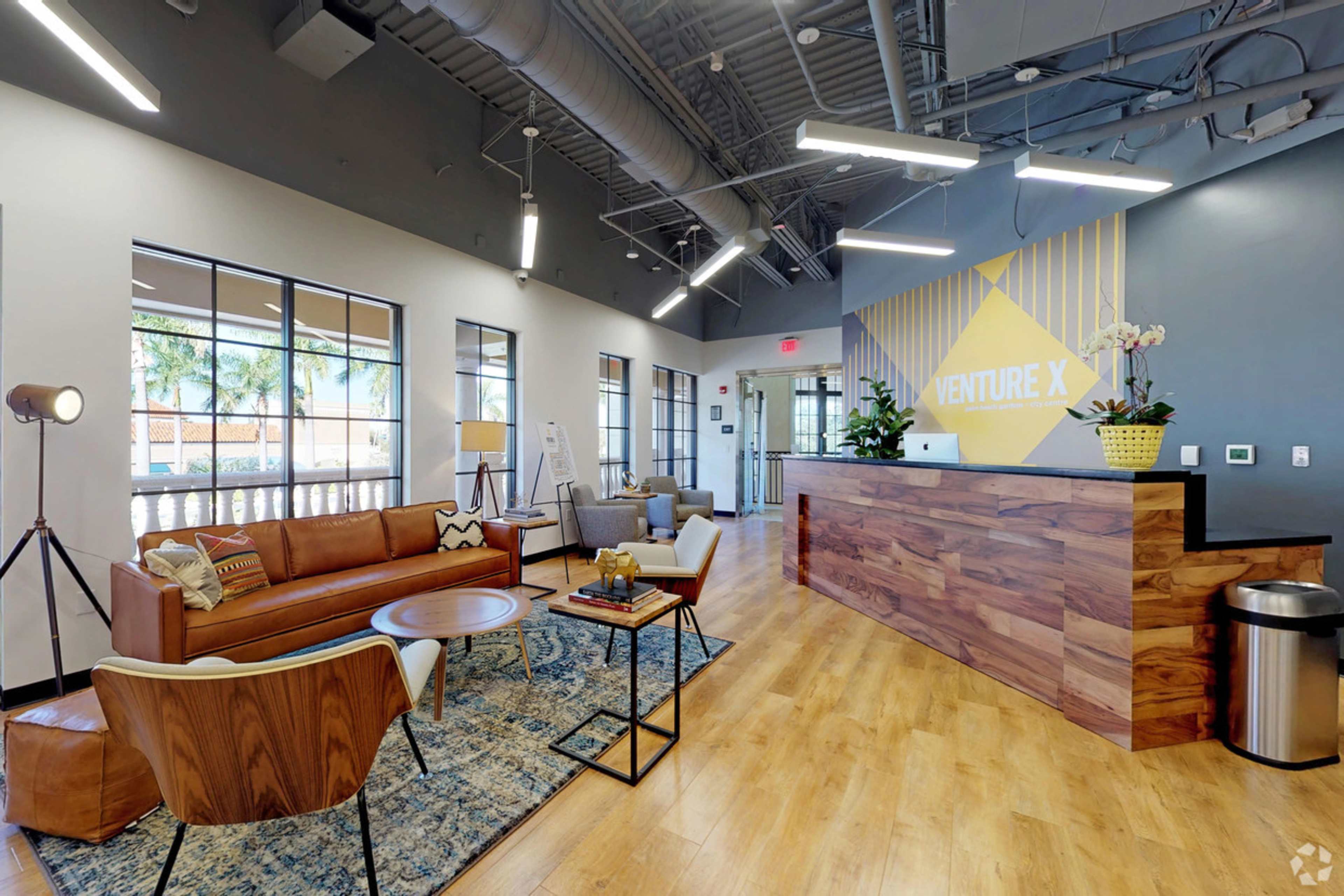A modern reception area featuring wooden furniture, a large desk with the text "VENTURE X," and a seating arrangement with various chairs and a coffee table.