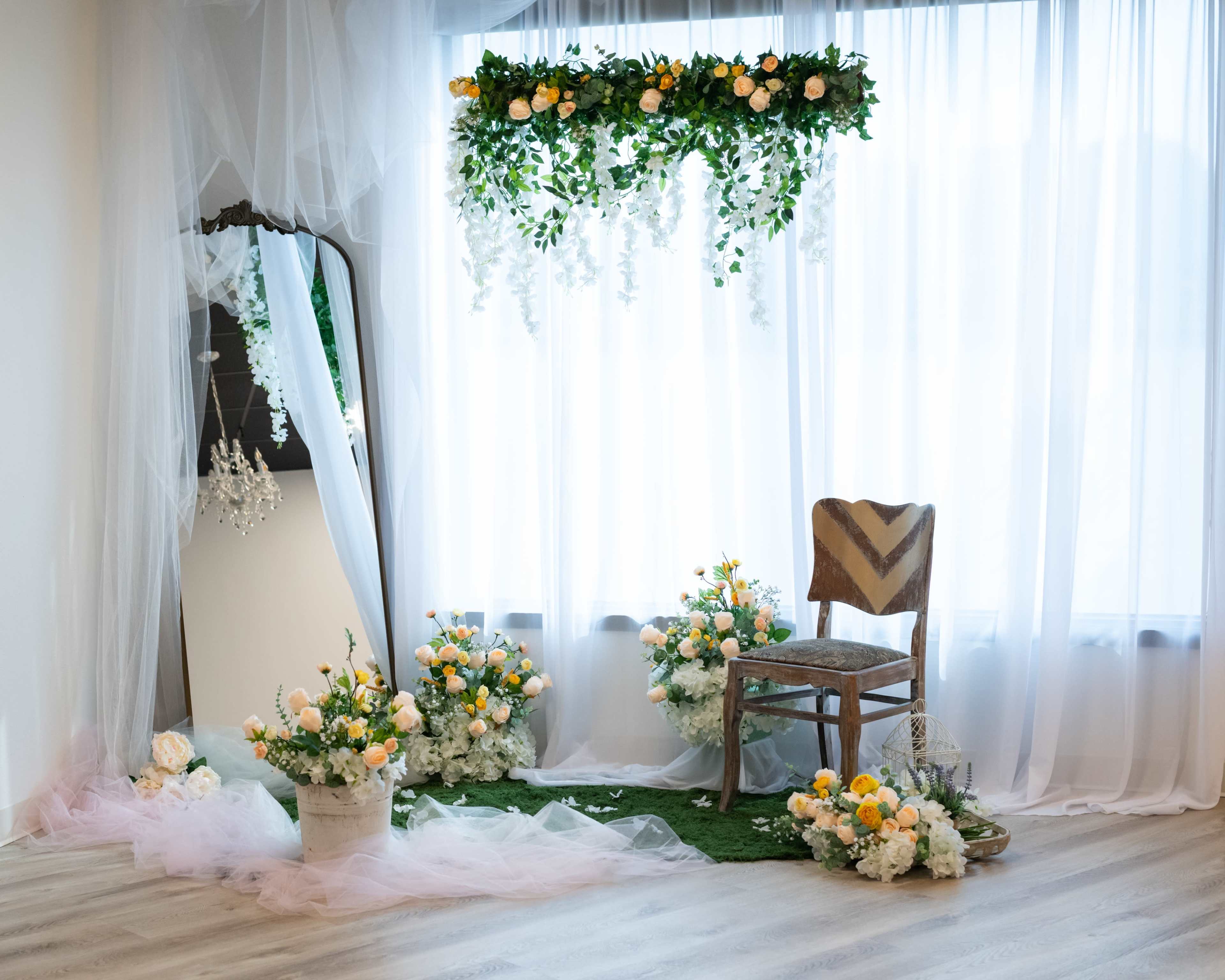 The image shows a decorated corner with a chair surrounded by floral arrangements, draped curtains, and a mirror, all set against a bright window.