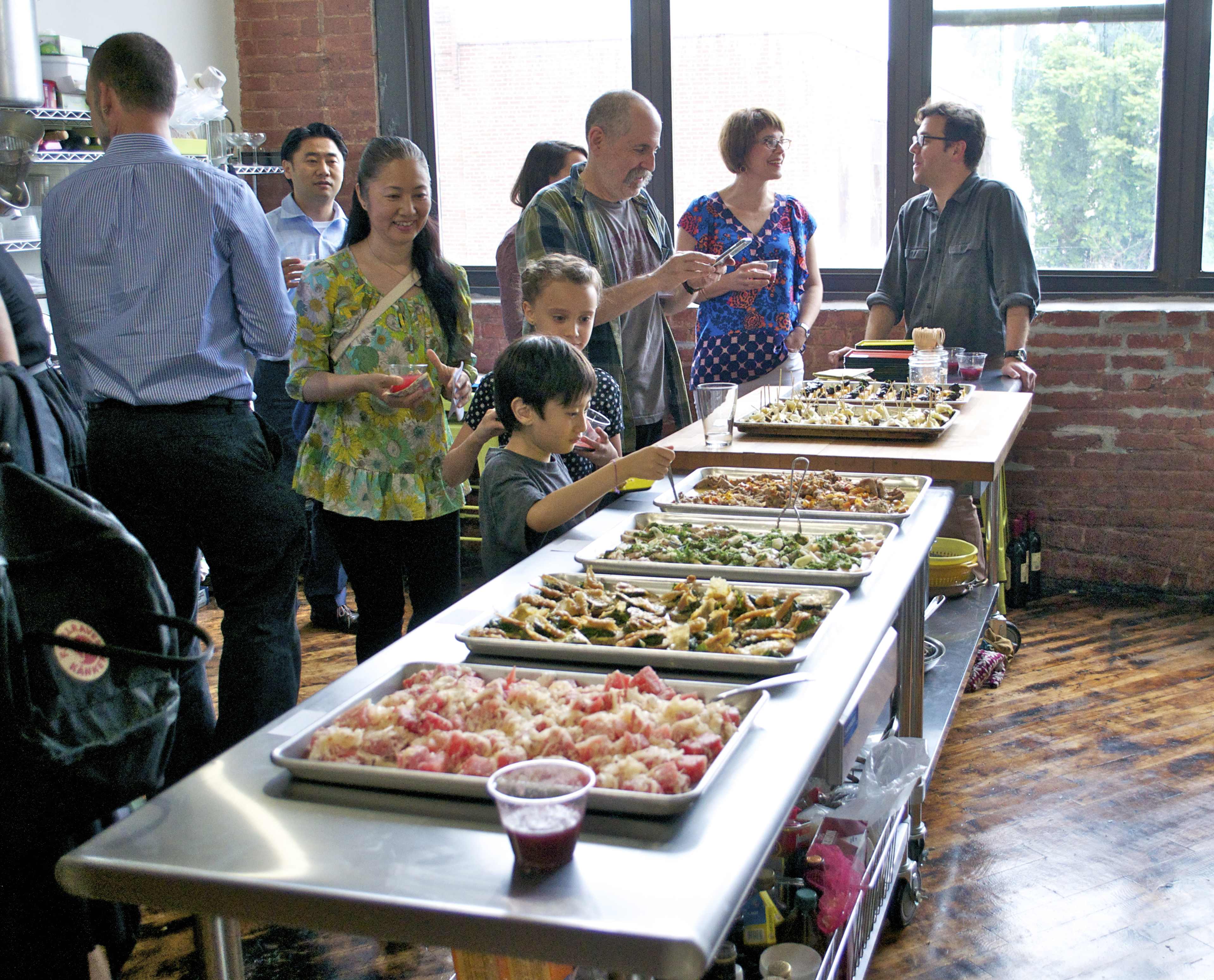 A group of people gathers around a table filled with various food platters in a casual indoor setting.