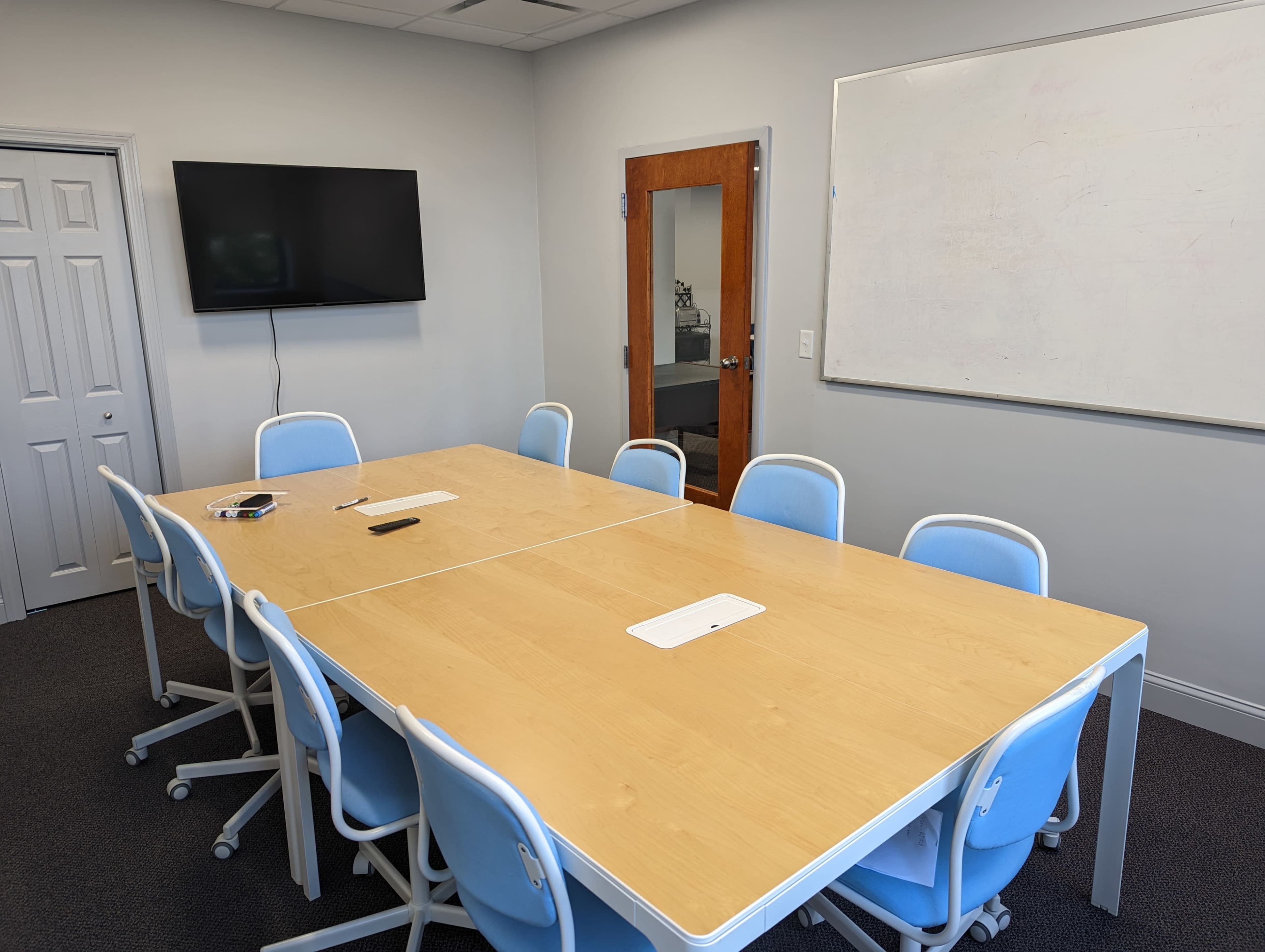 A long wooden conference table with ten blue chairs is positioned in a meeting room, featuring a television on the wall and a whiteboard nearby.