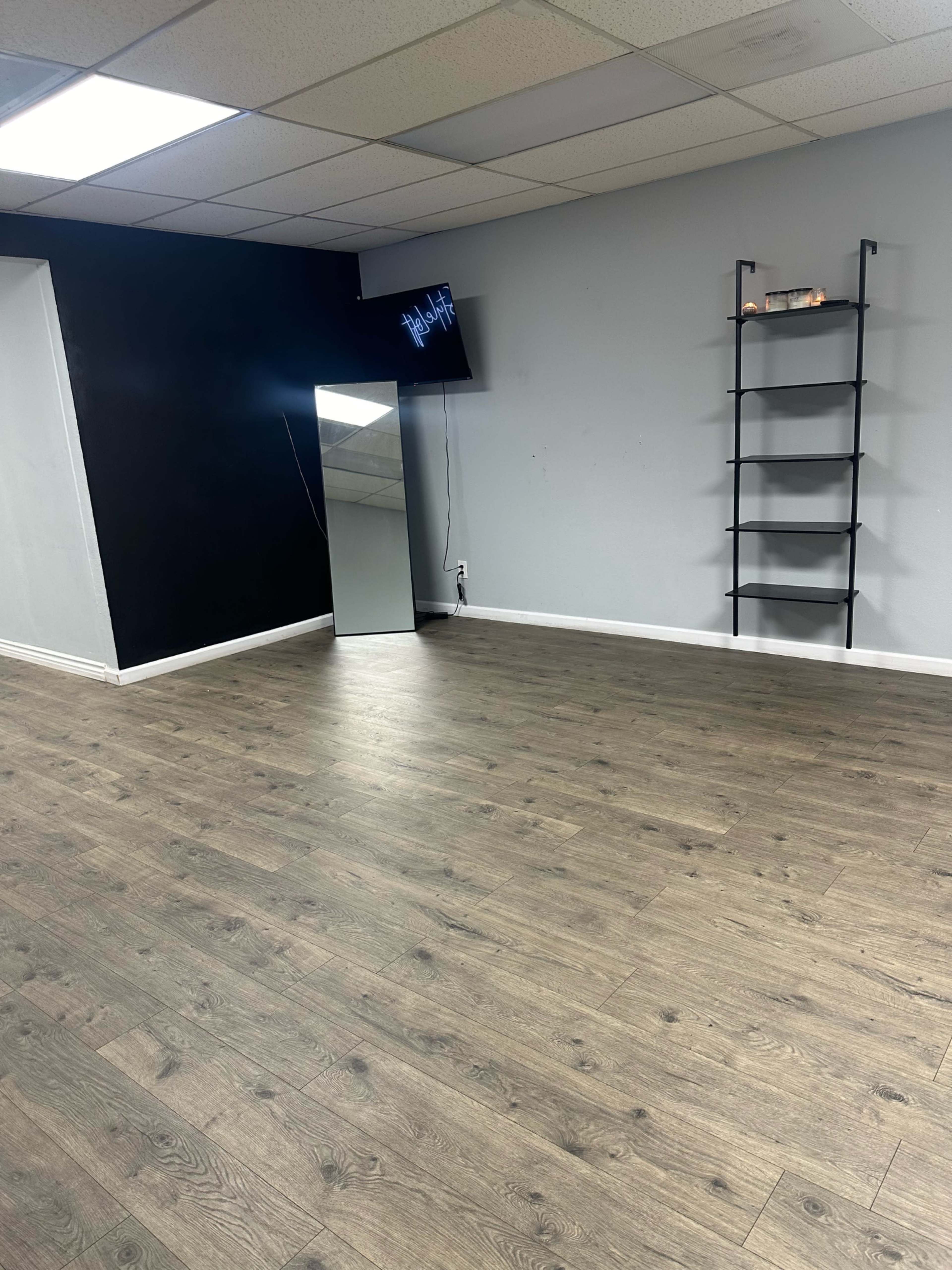 The image shows a spacious, empty room with a wooden floor, a wall-mounted TV, a tall shelf, and a large mirror along one wall.