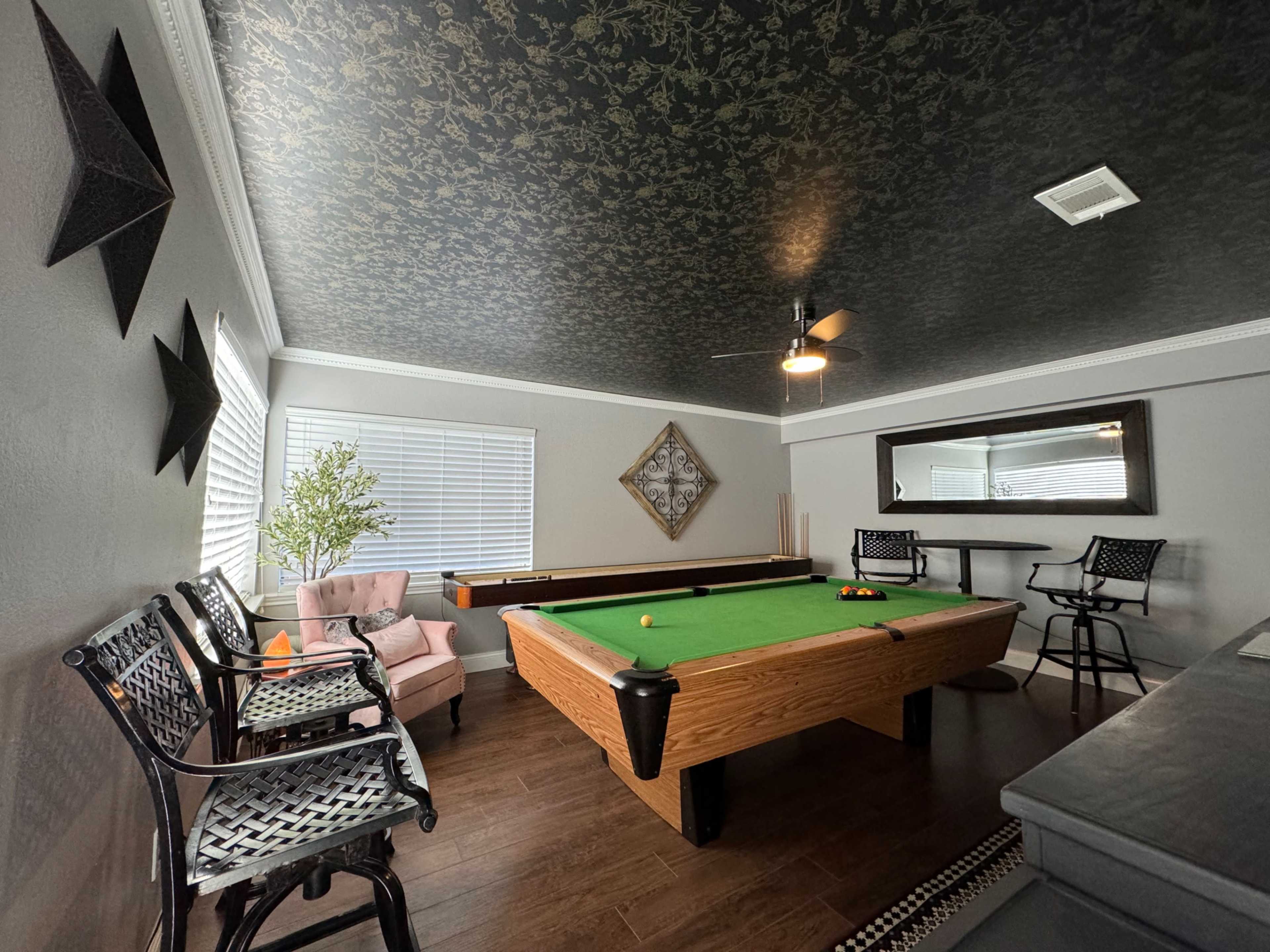 A game room featuring a green pool table, seating areas with a pink armchair and bar stools, and a black decorative ceiling.