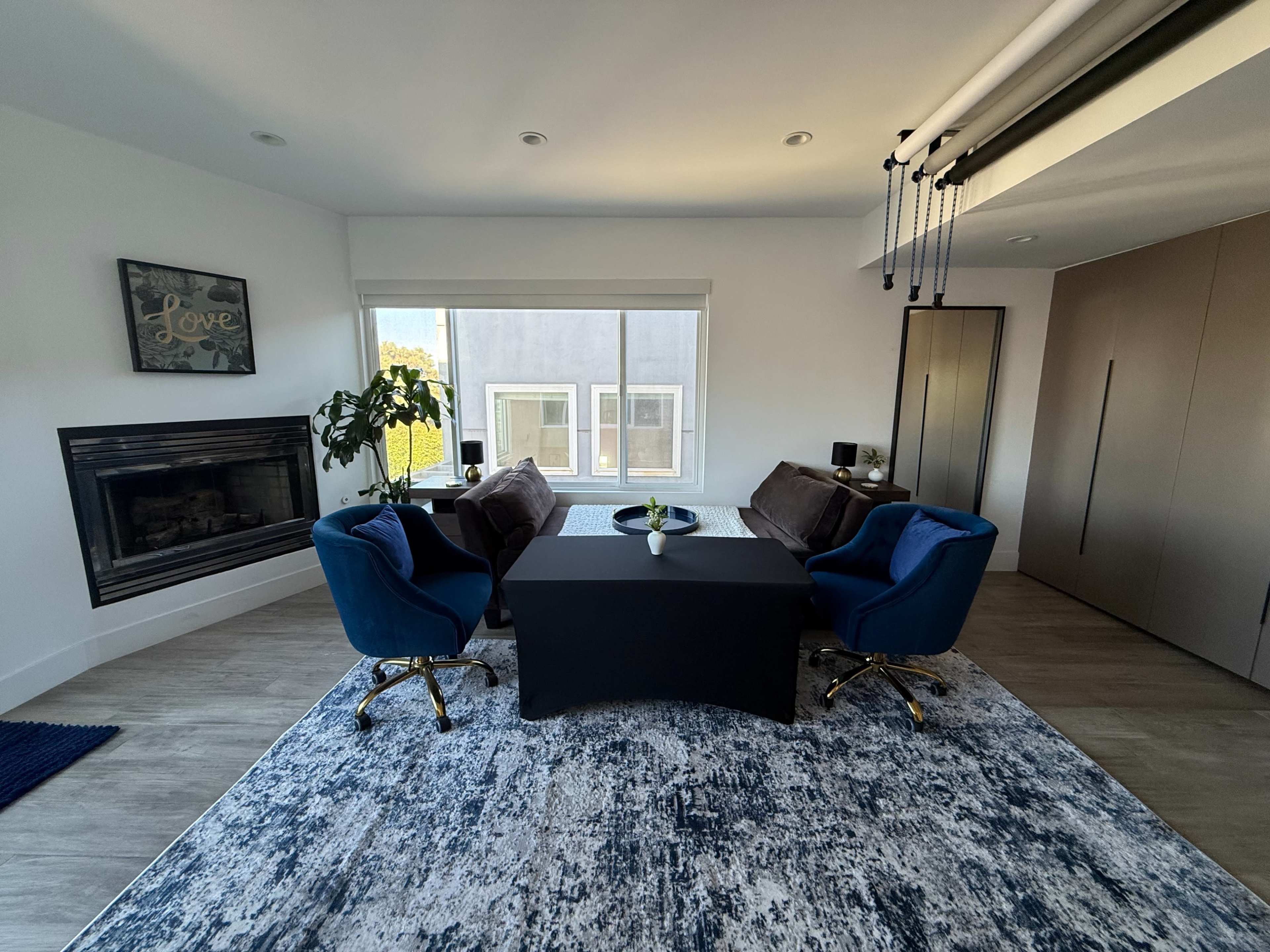 The image shows a modern room with two blue chairs and a black table placed on a patterned rug, featuring a fireplace and large windows.