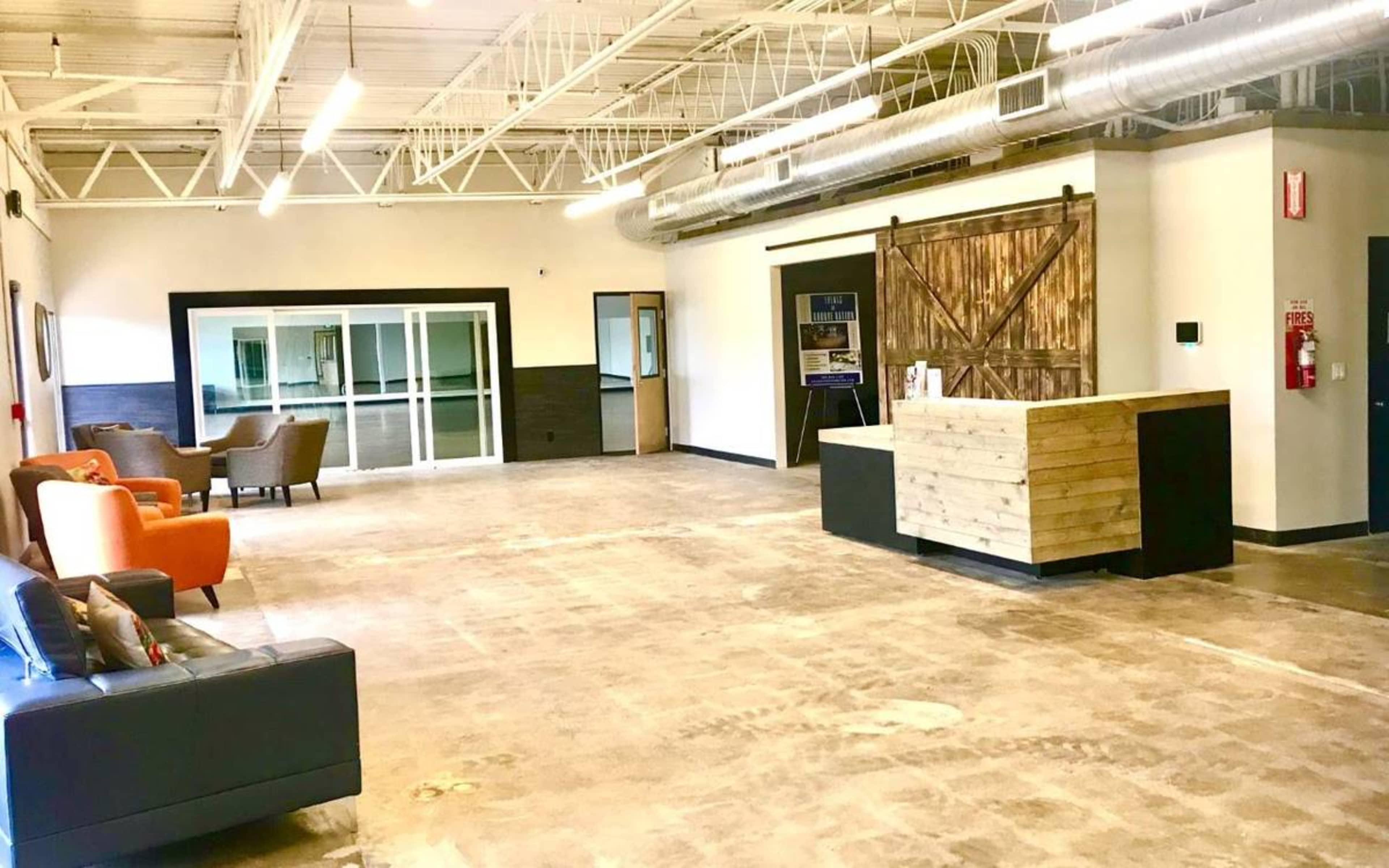 A spacious, unfinished reception area with a wooden front desk and a few seating options, including a black sofa and orange chairs.