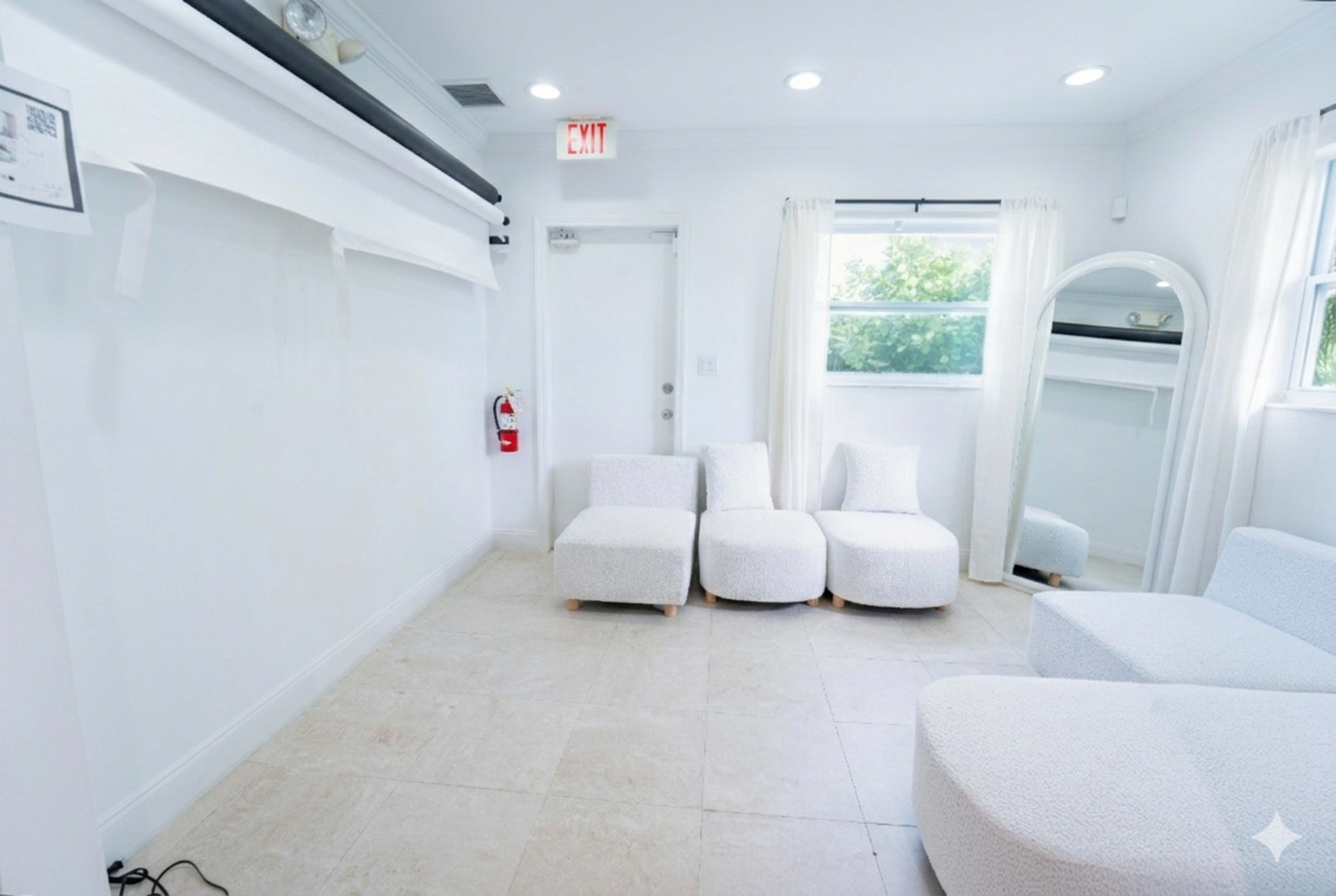 The image shows a bright, minimalist room with light-colored walls, a window, and three upholstered seating elements.