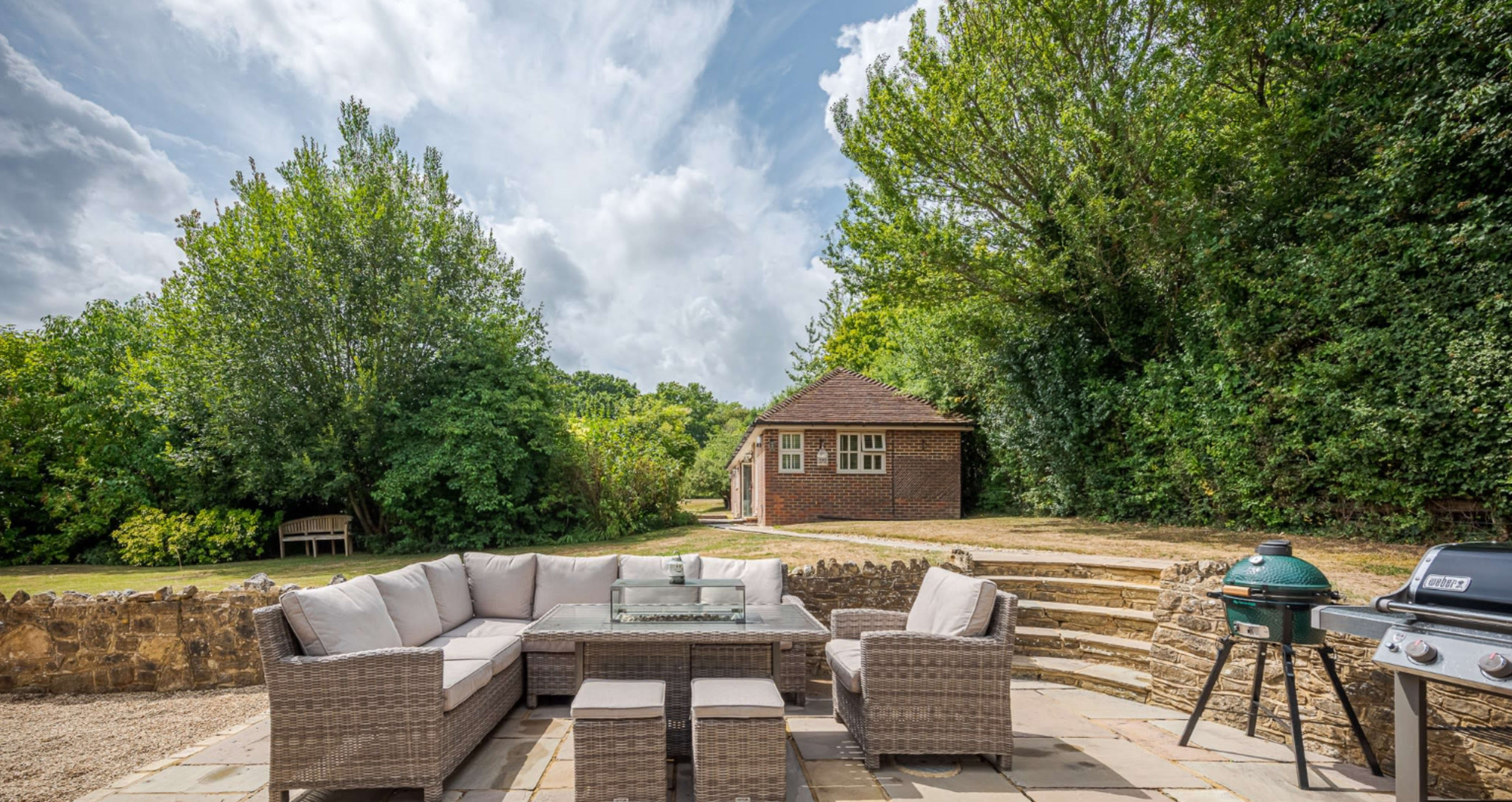 A patio area features a large seating arrangement with a glass table, surrounded by trees and a building in the background.