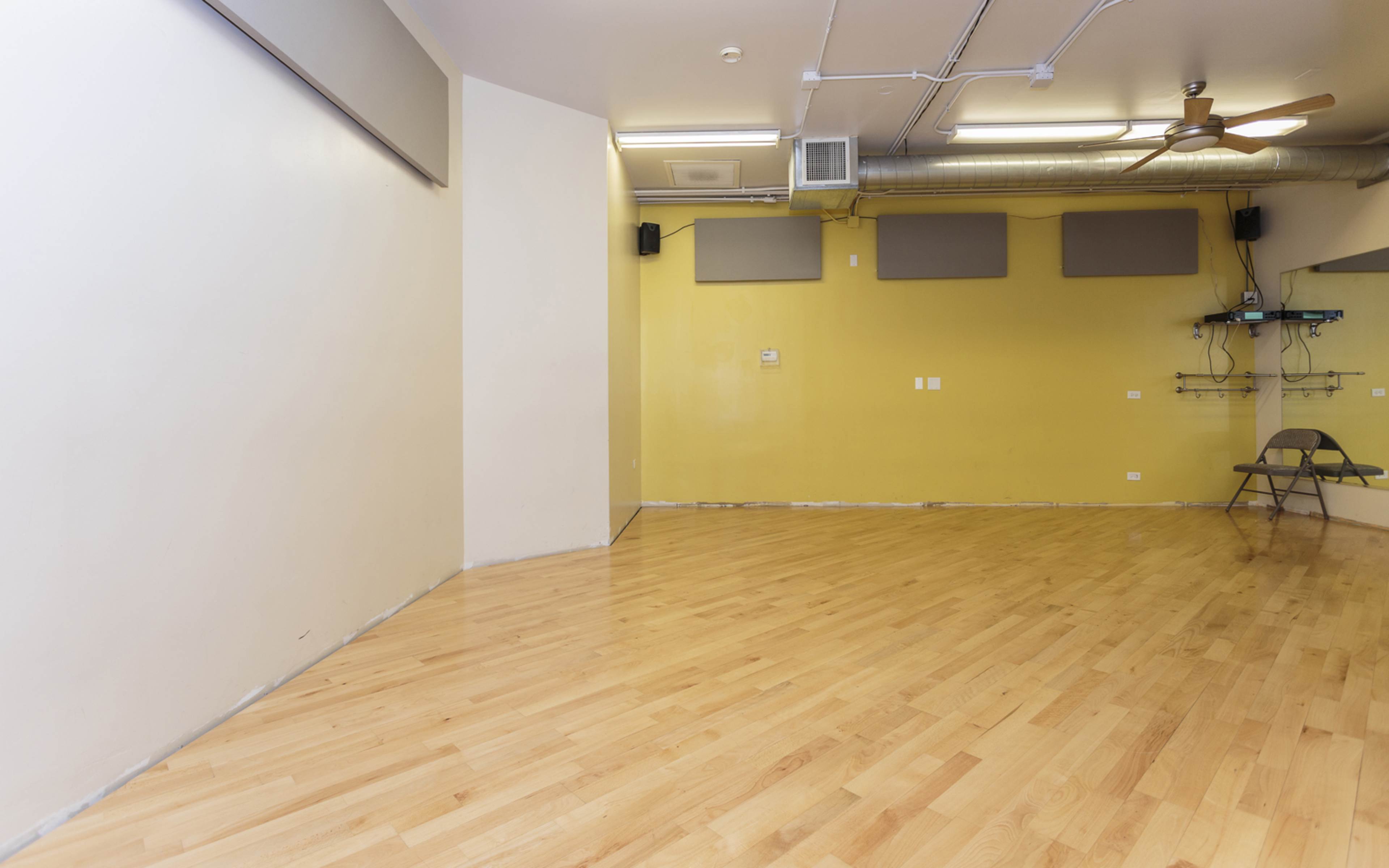The image shows an empty room with wooden flooring, yellow walls, and minimal equipment mounted on the walls.