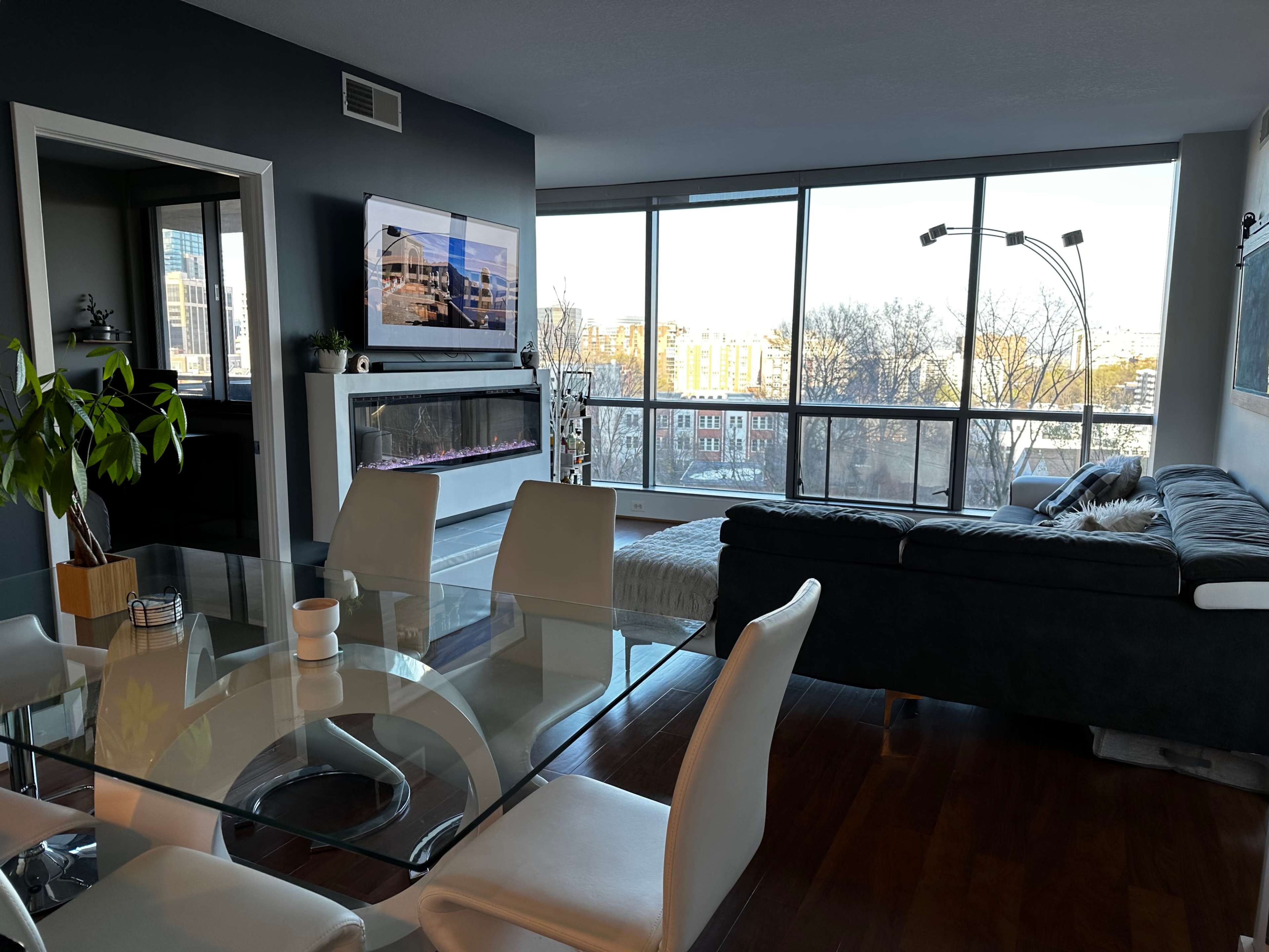 A modern living space with a dining area featuring a glass table and white chairs, a dark gray couch, and large windows offering a view of the city.