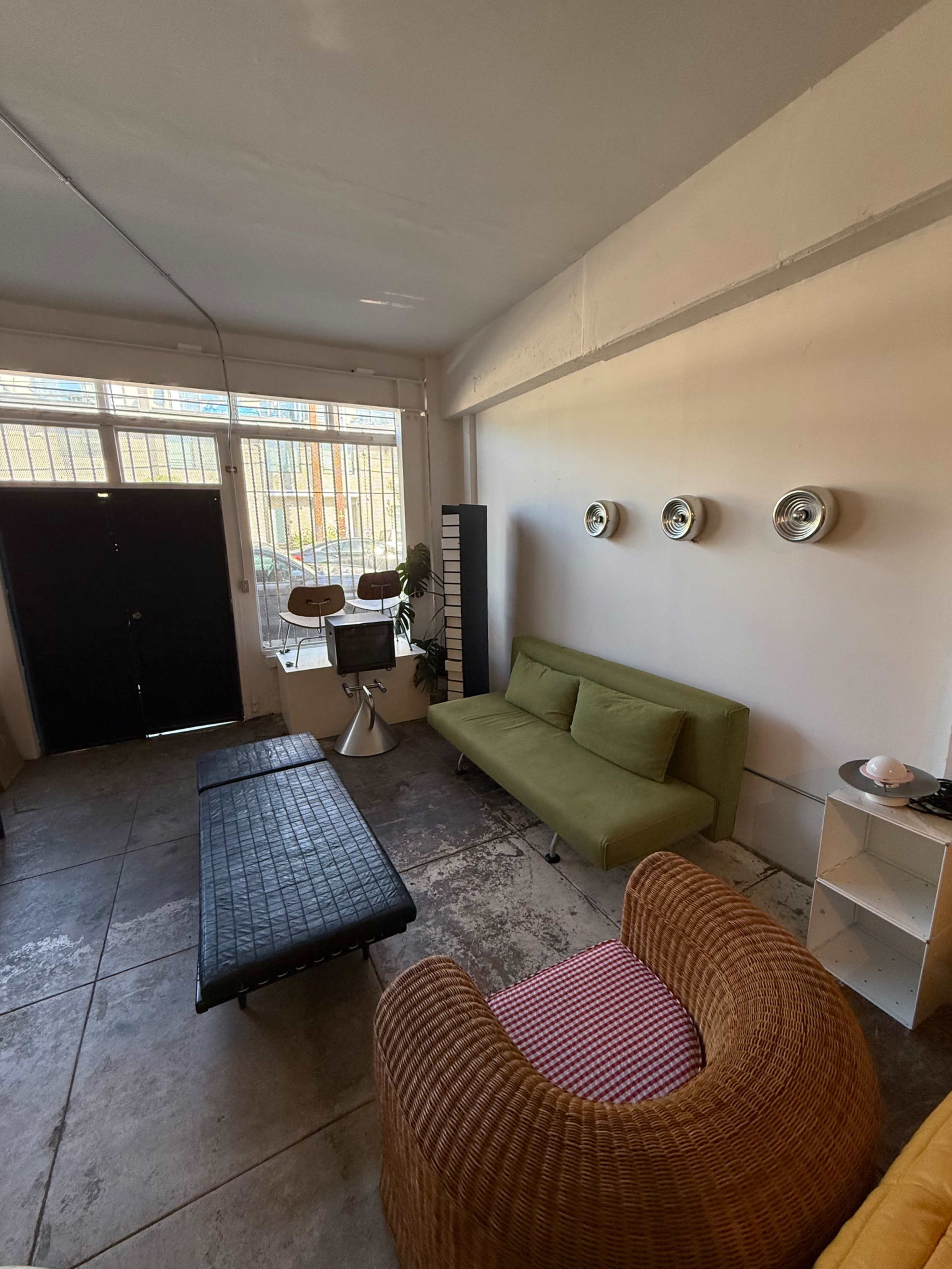 The image shows a modern, minimalist interior with a green sofa, a wicker chair, and a black bench on a tiled floor, illuminated by natural light from a large window.