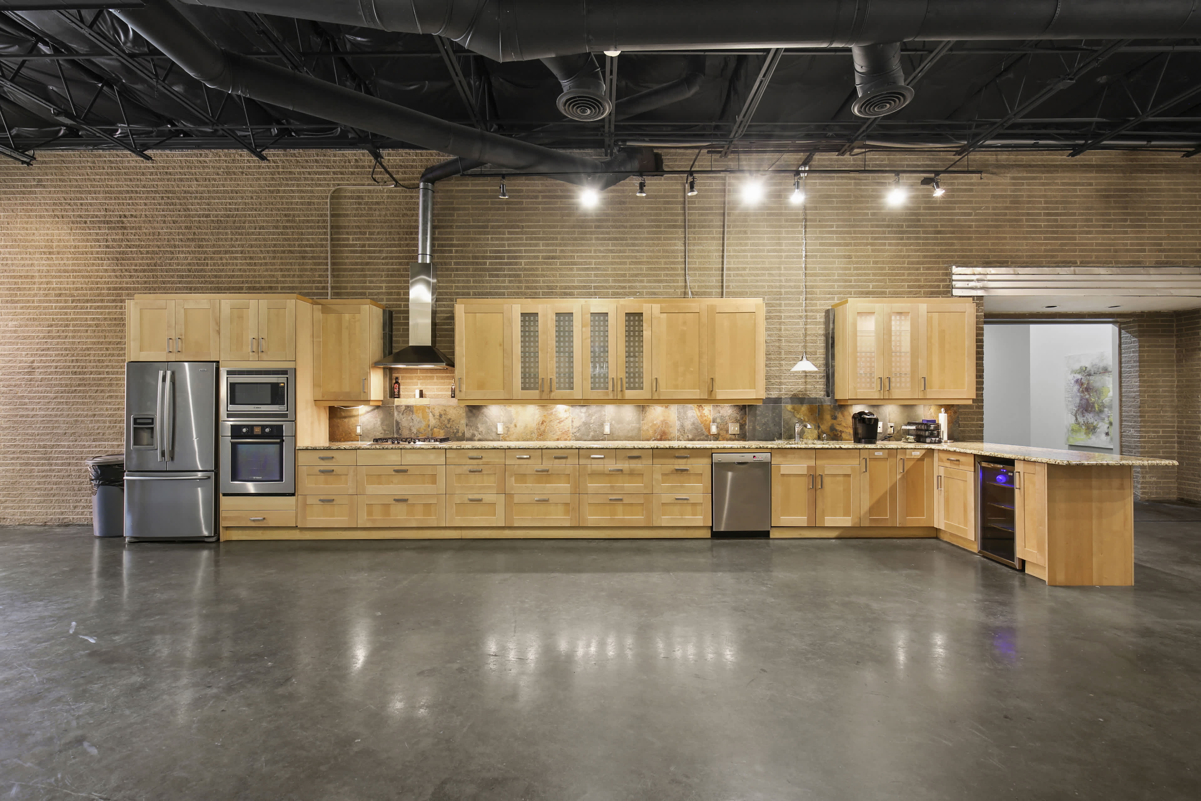 The image shows a large kitchen with wooden cabinetry, stainless steel appliances, and a granite countertop in a modern industrial space.