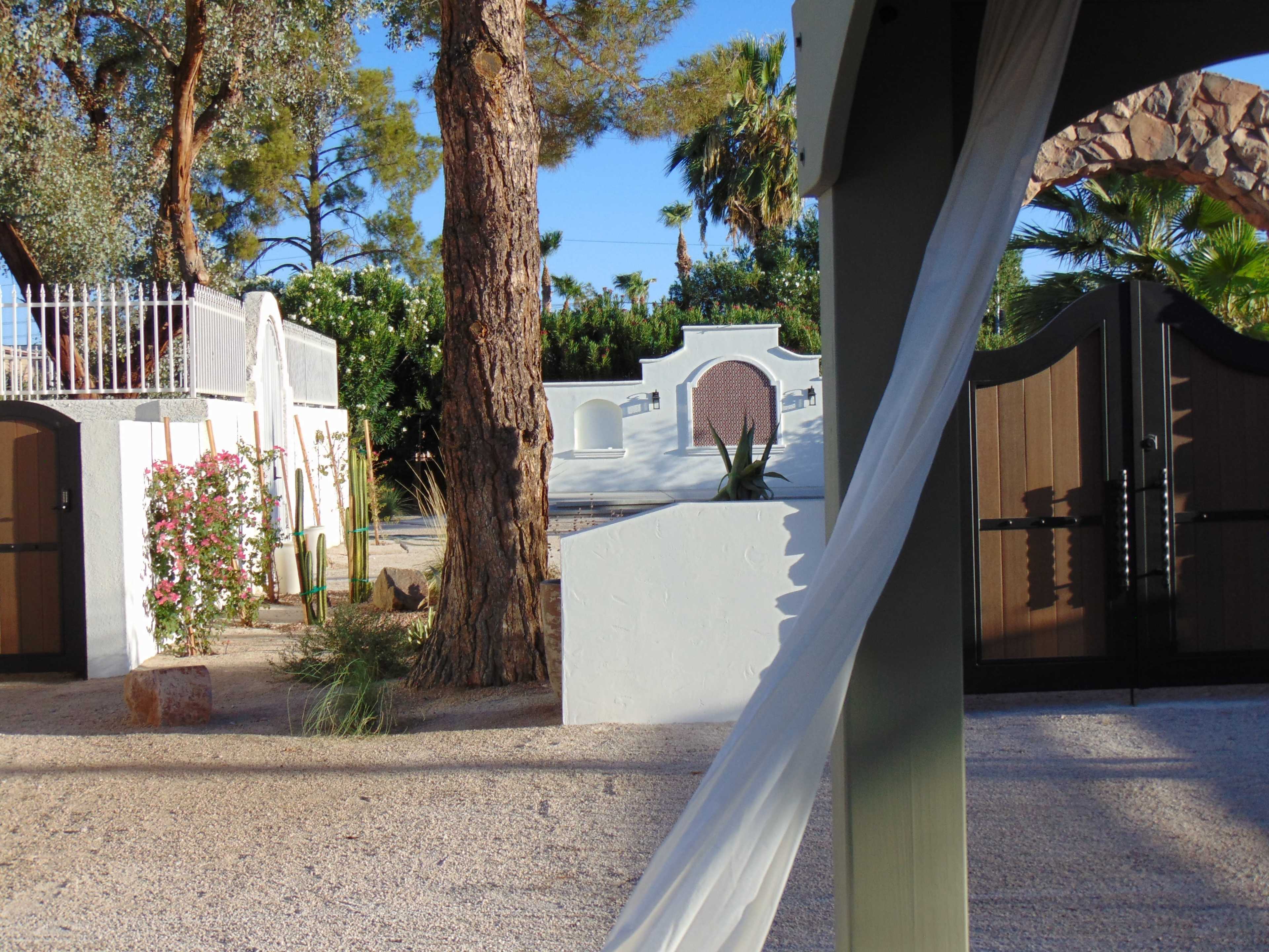 The image shows a tranquil outdoor scene featuring a gravel pathway lined with sparse vegetation, flanked by white buildings and wooden gates surrounded by palm trees.