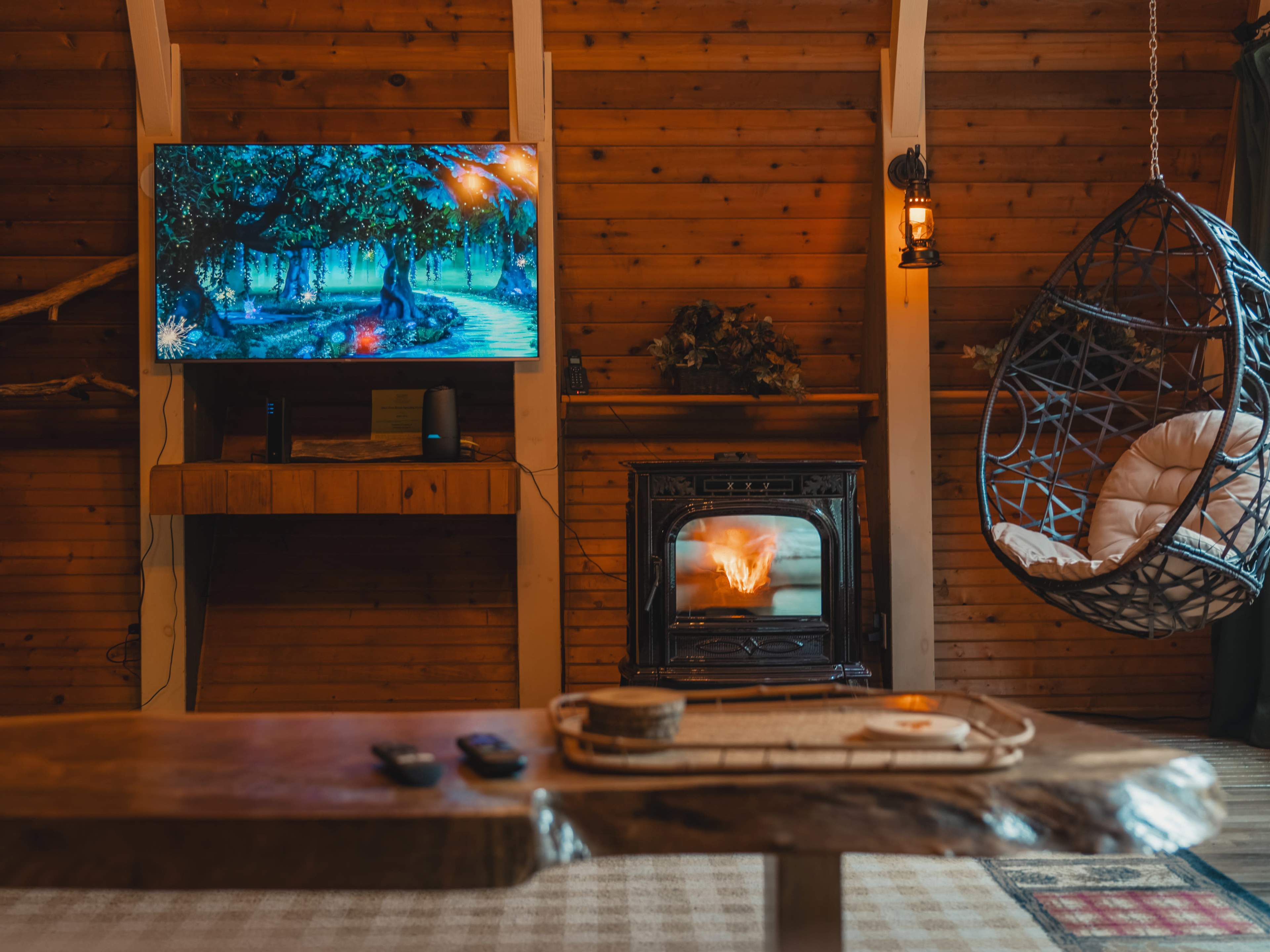 A wooden cabin interior featuring a television mounted on the wall, a hanging chair, a wood stove with a visible fire, and a rustic wooden coffee table.