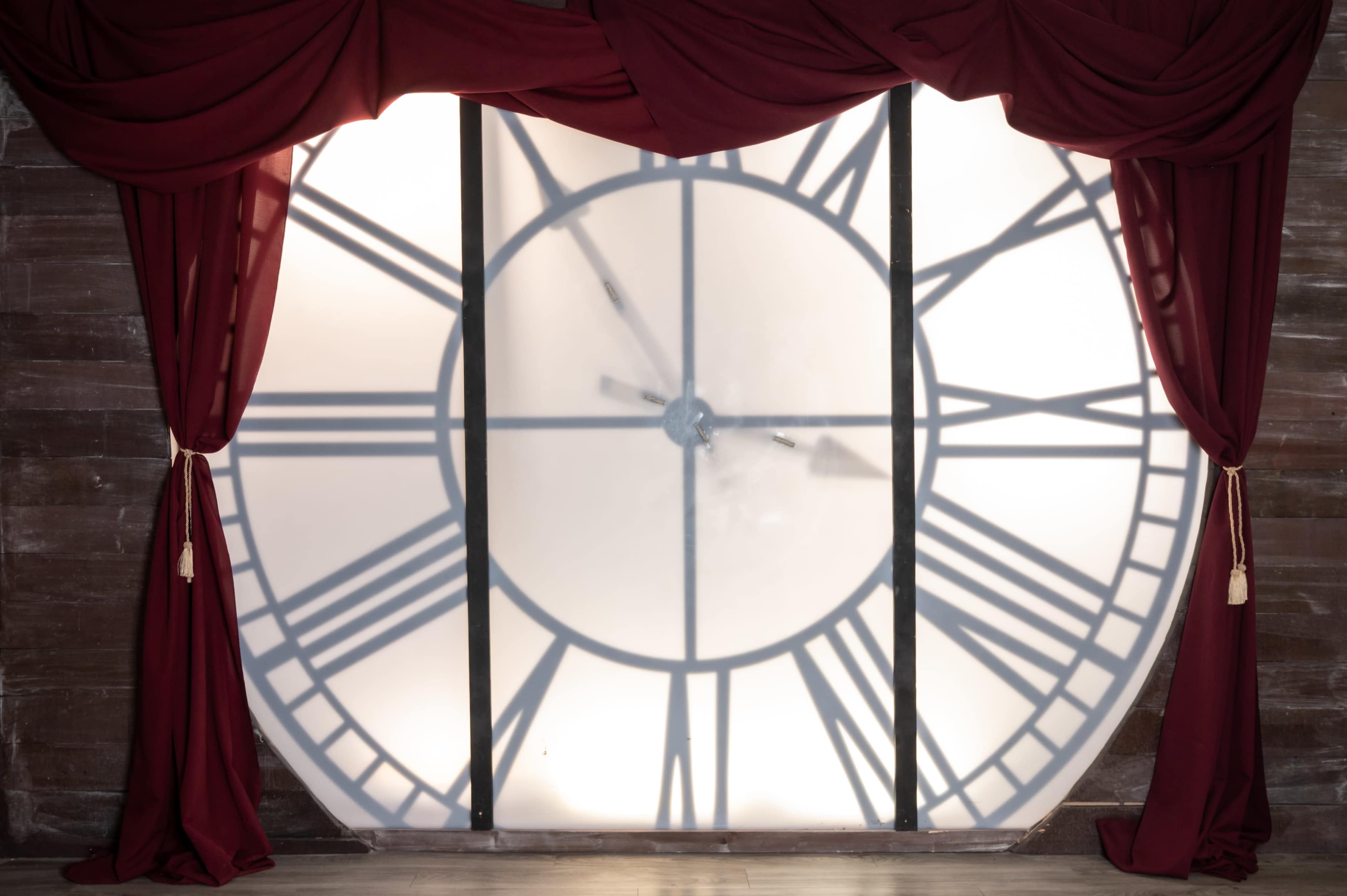 A large clock face with Roman numerals is partially obscured by sheer curtains, creating a silhouette effect.