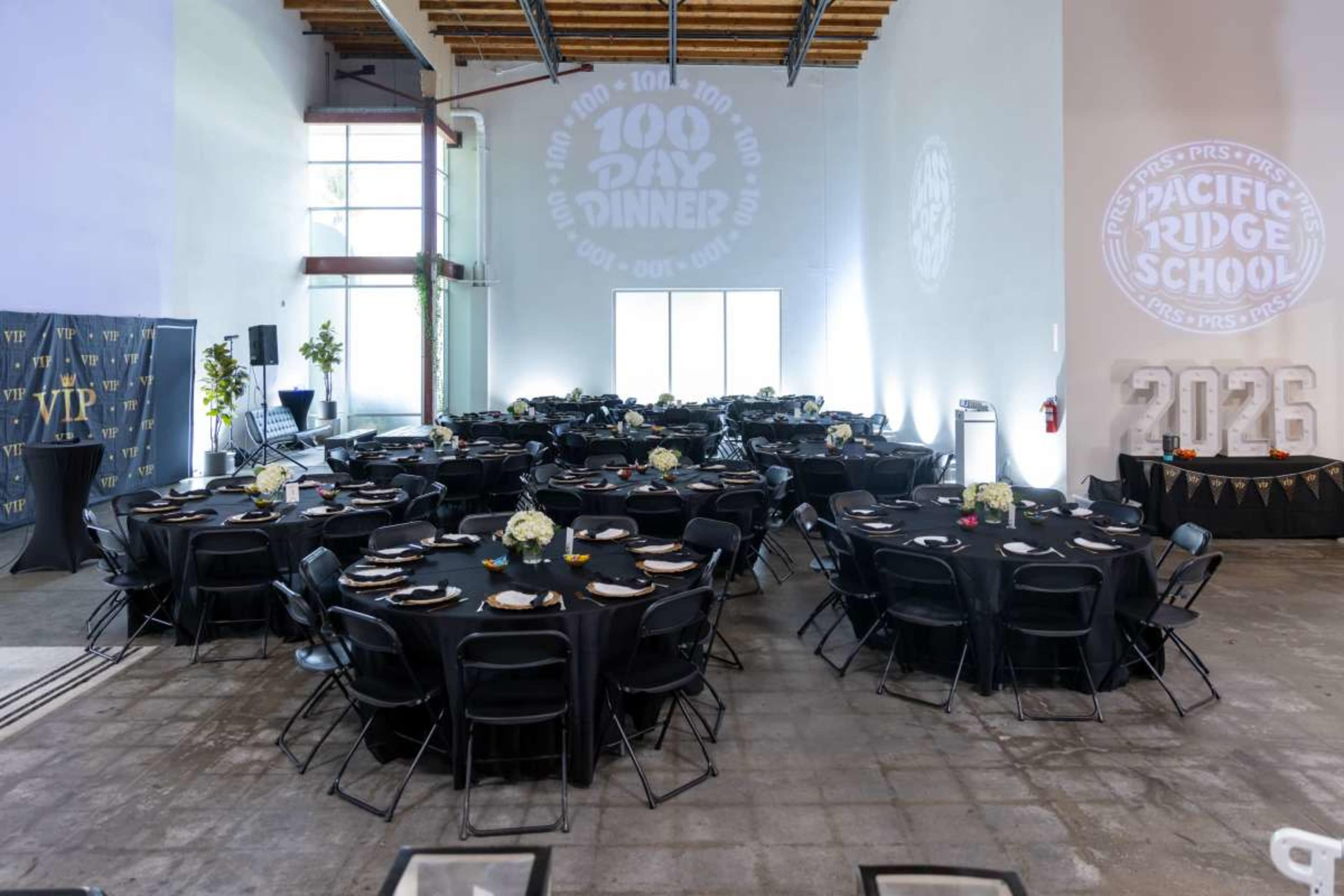 The image shows a spacious event hall set up with round tables covered in black tablecloths, arranged for a dinner event, with a projection on the wall displaying "100 Day Dinner" and "Pacific Ridge School."