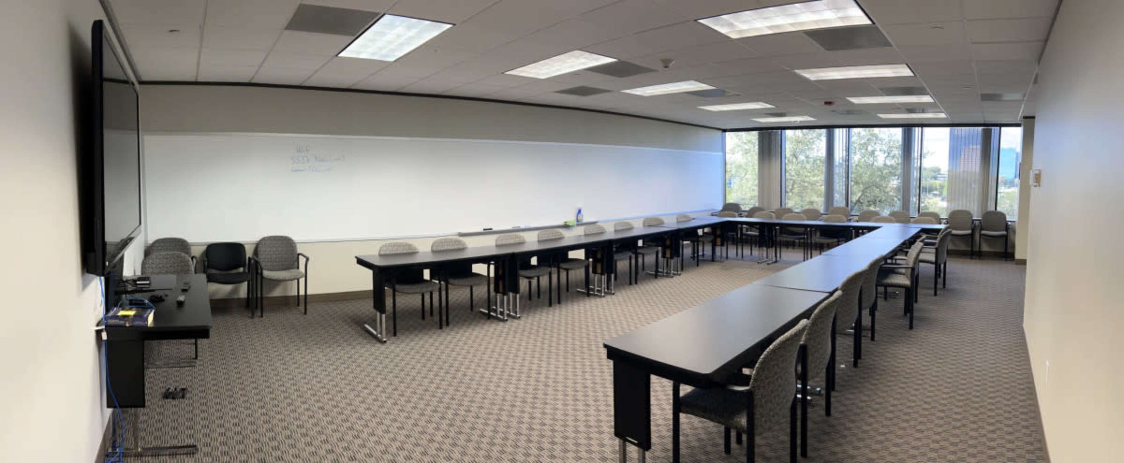 A conference room with multiple long tables arranged in a U-shape and chairs along the sides, surrounded by large windows.