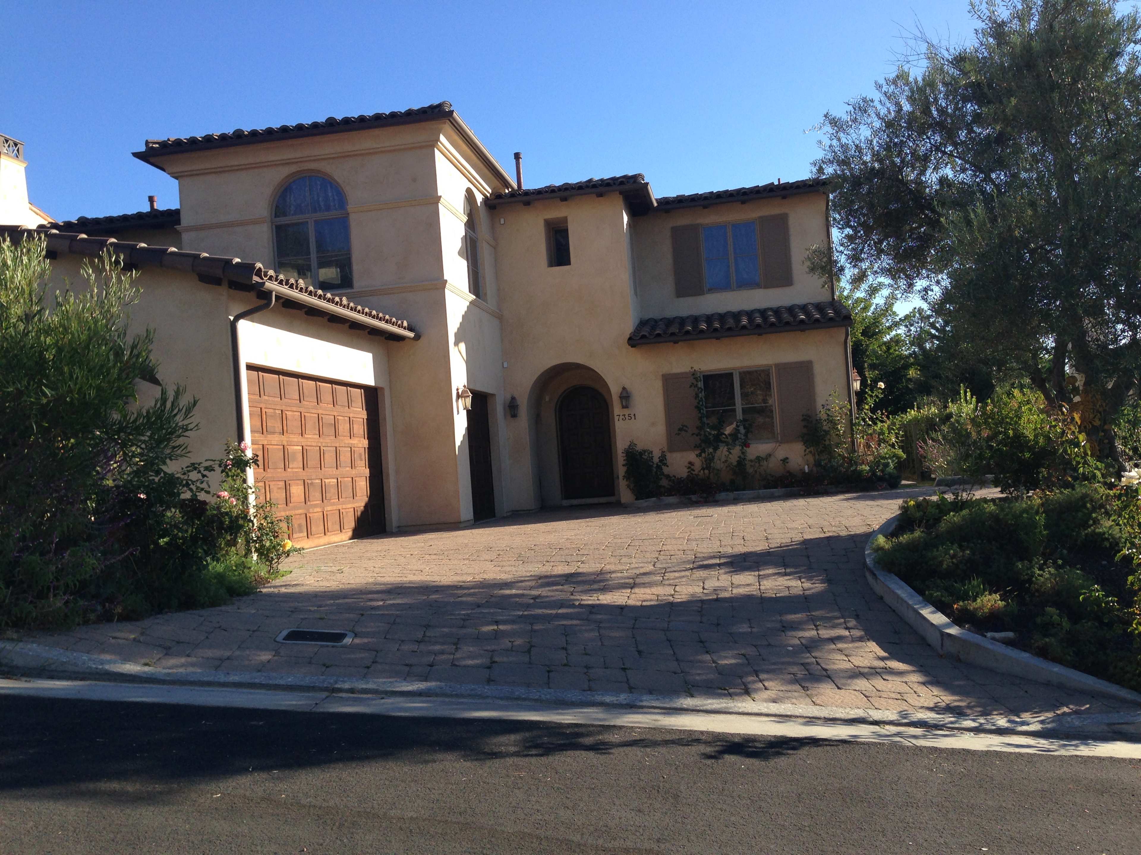 The image shows a two-story, Mediterranean-style house with a tiled roof, a stone driveway, and landscaping along the front.