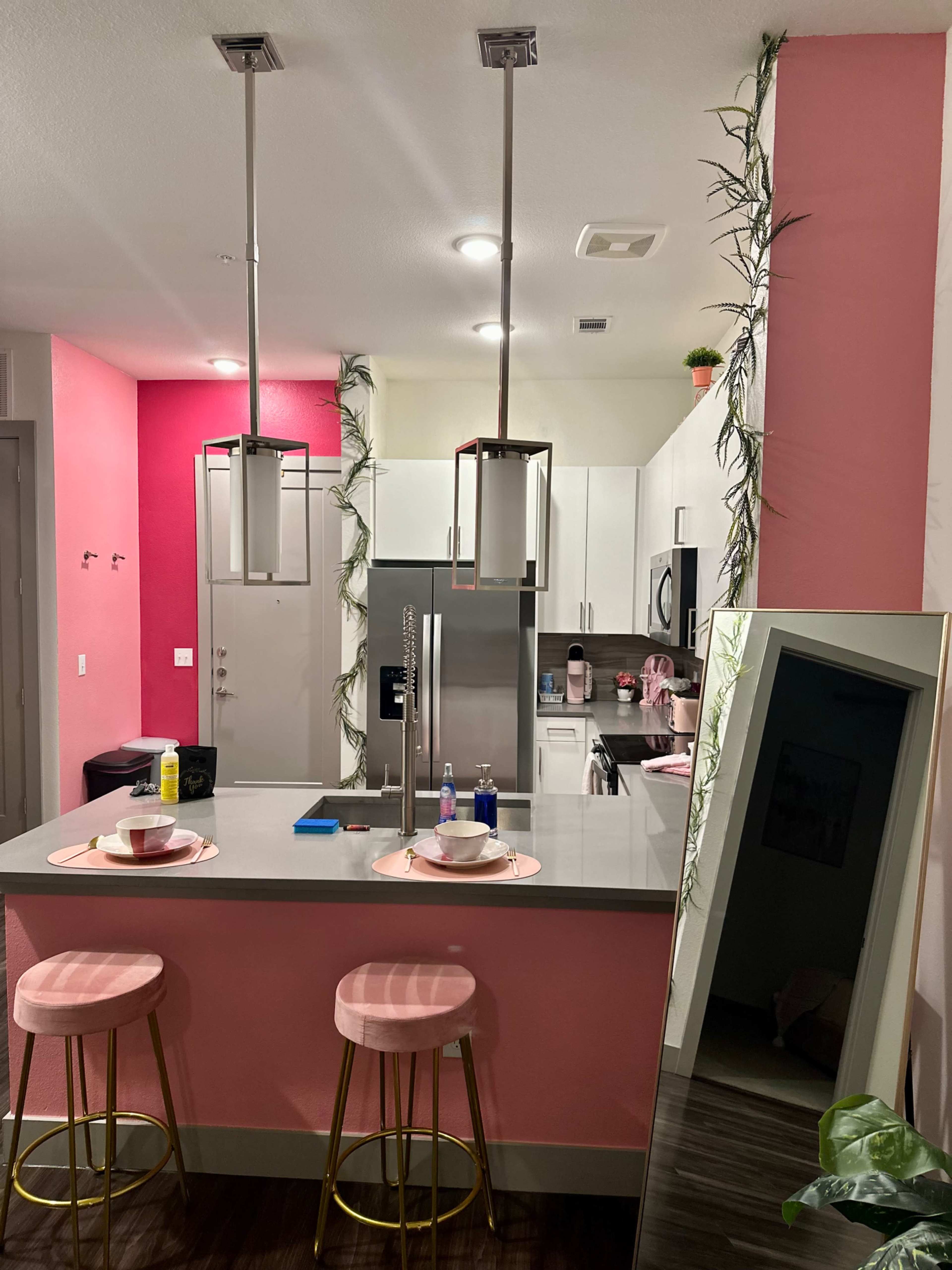 A modern kitchen area with pink accent walls, bar stools, and a mirror reflecting the space.