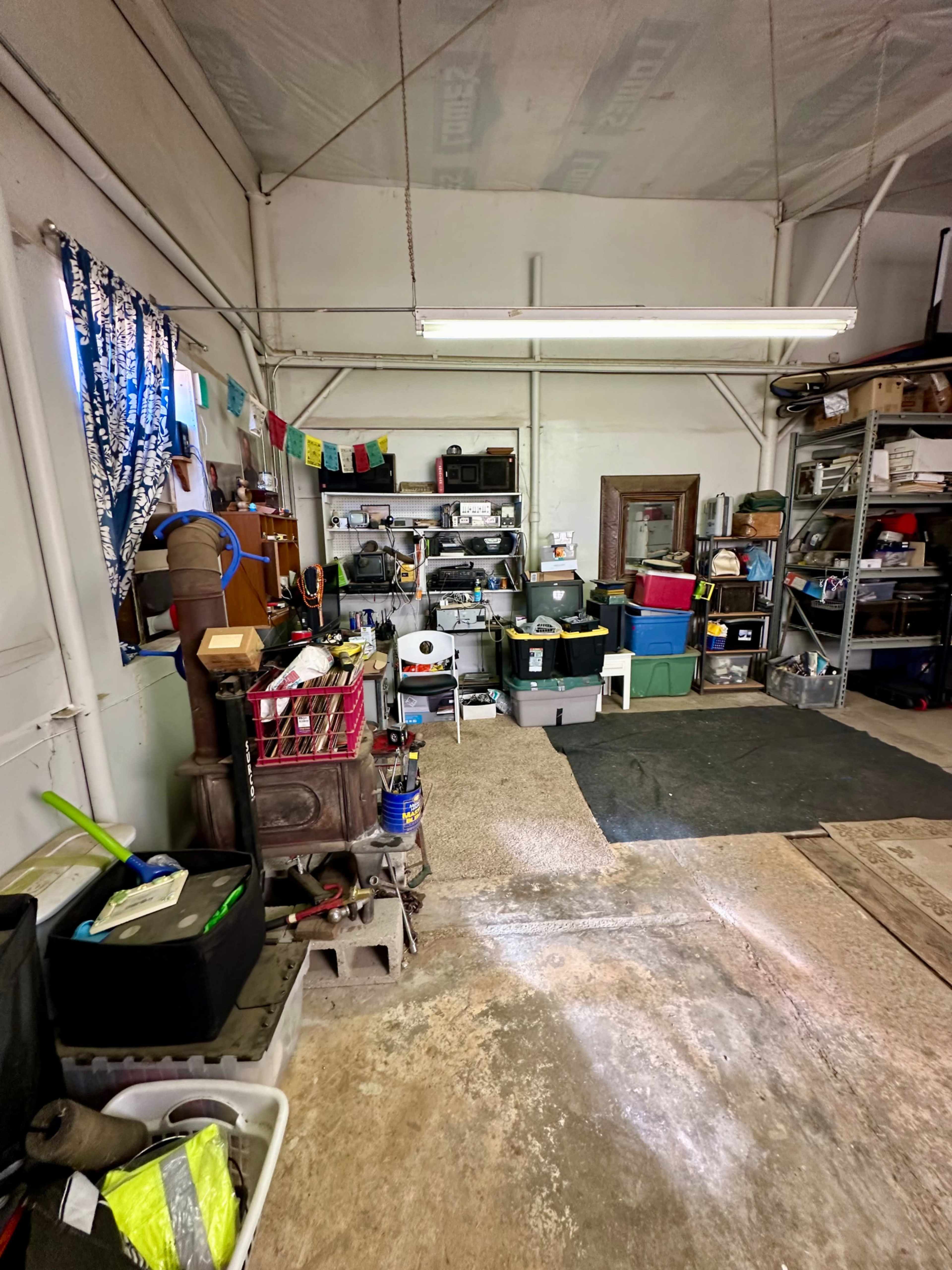 The image shows a cluttered garage or workshop space with various storage bins, tools, and a wood stove along one side.