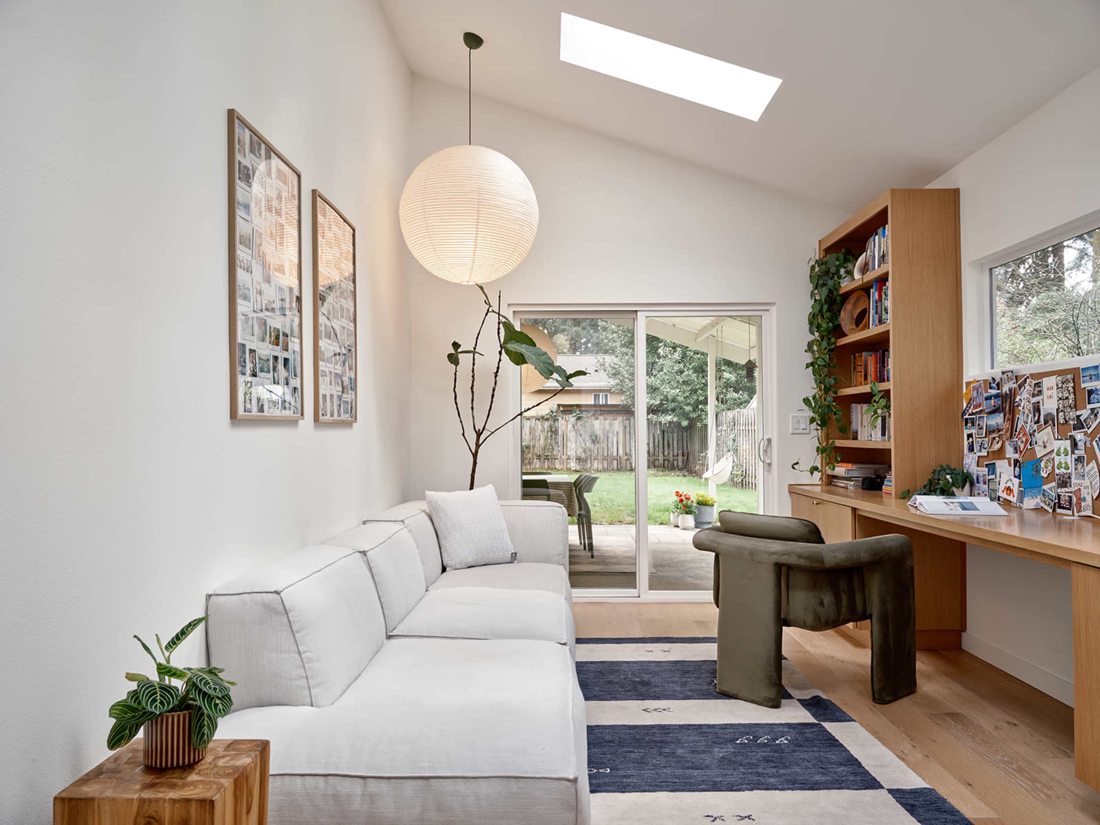 The image shows a modern living room with a light-colored sectional sofa, a large paper lantern, and a workspace with a bookshelf, overlooking a backyard through sliding glass doors.