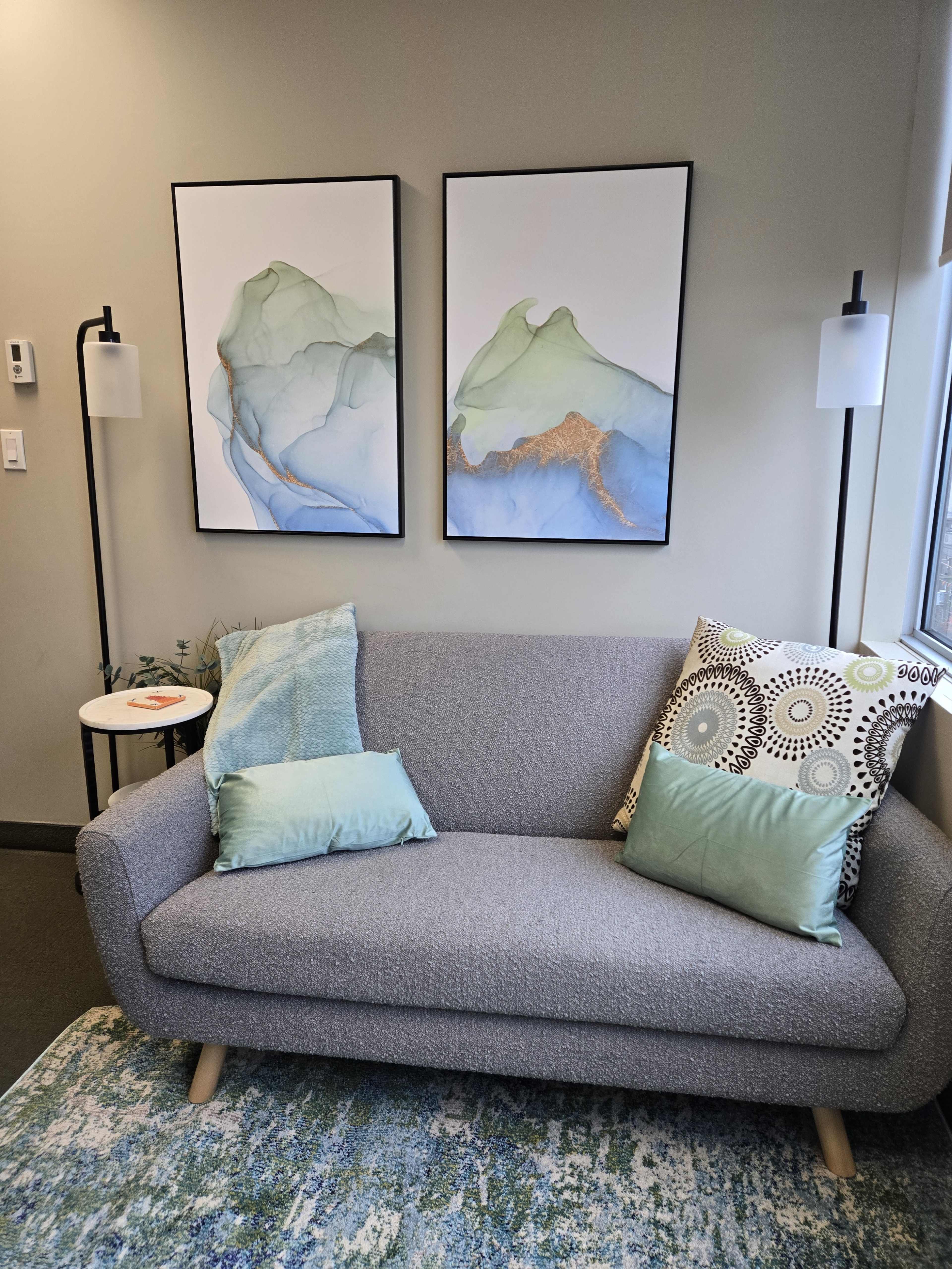 The image shows a gray sofa adorned with two light blue pillows and a decorative patterned pillow, positioned between two lamps and beneath two framed abstract art pieces on a wall.